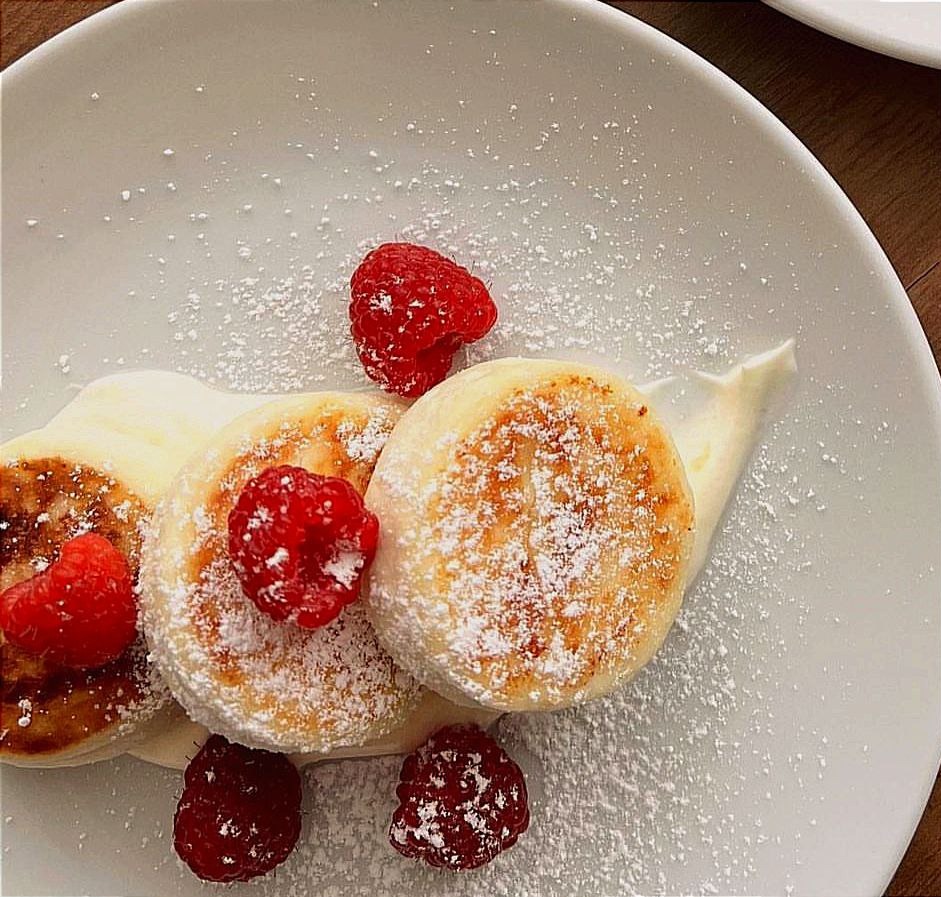 Trzy okrągłe serniczki polane śmietanką i posypane cukrem pudrem, udekorowane świeżymi malinami na białym talerzu, widok z góry.