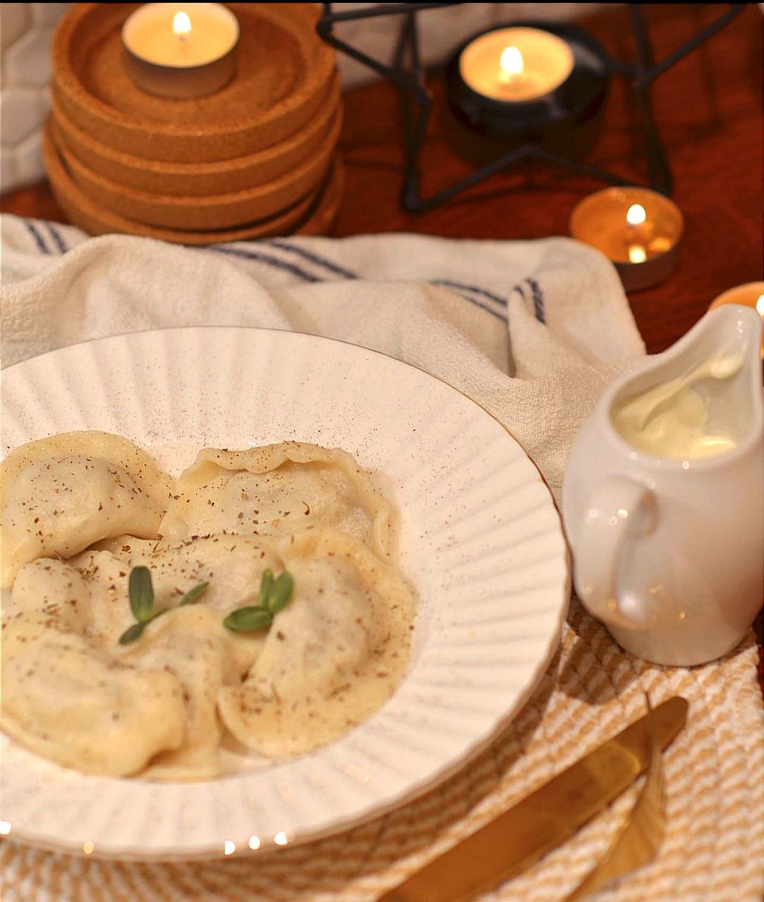 Świąteczne pierogi na białym talerzu z sosem śmietanowym, dekorowane świeżymi ziołami i przyprawami, w tle świece na drewnianym stole, tworzące ciepłą, świąteczną atmosferę.