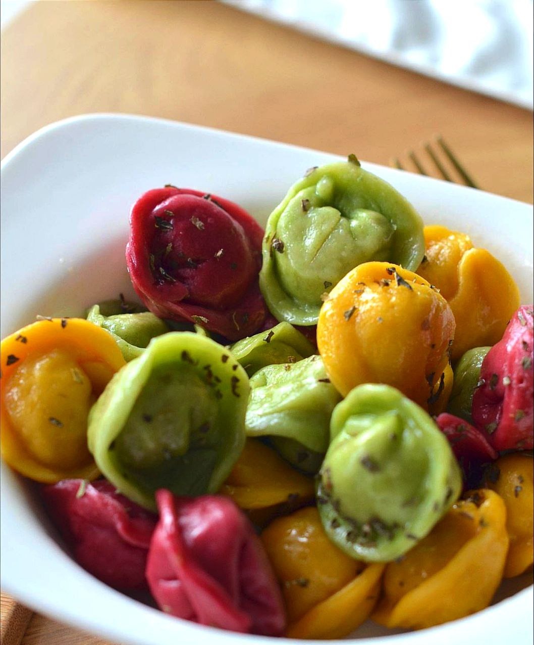 Ręcznie robione, kolorowe pierożki w białej miseczce, posypane ziołami, na drewnianym stole.