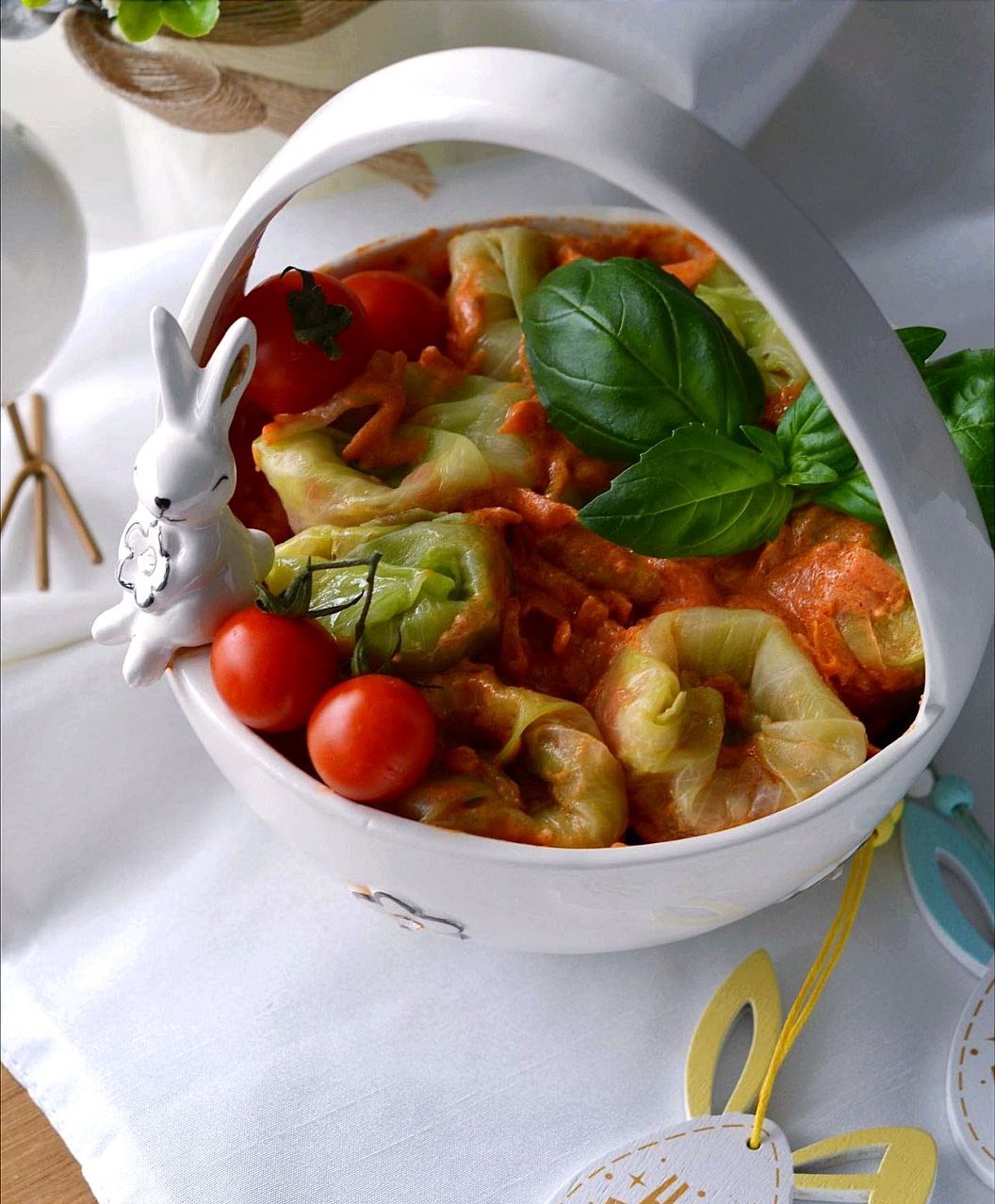 Gołąbki w sosie pomidorowym, podane w białym koszyczku z ceramicznym zajączkiem i dekoracjami wielkanocnymi, na białym obrusie.