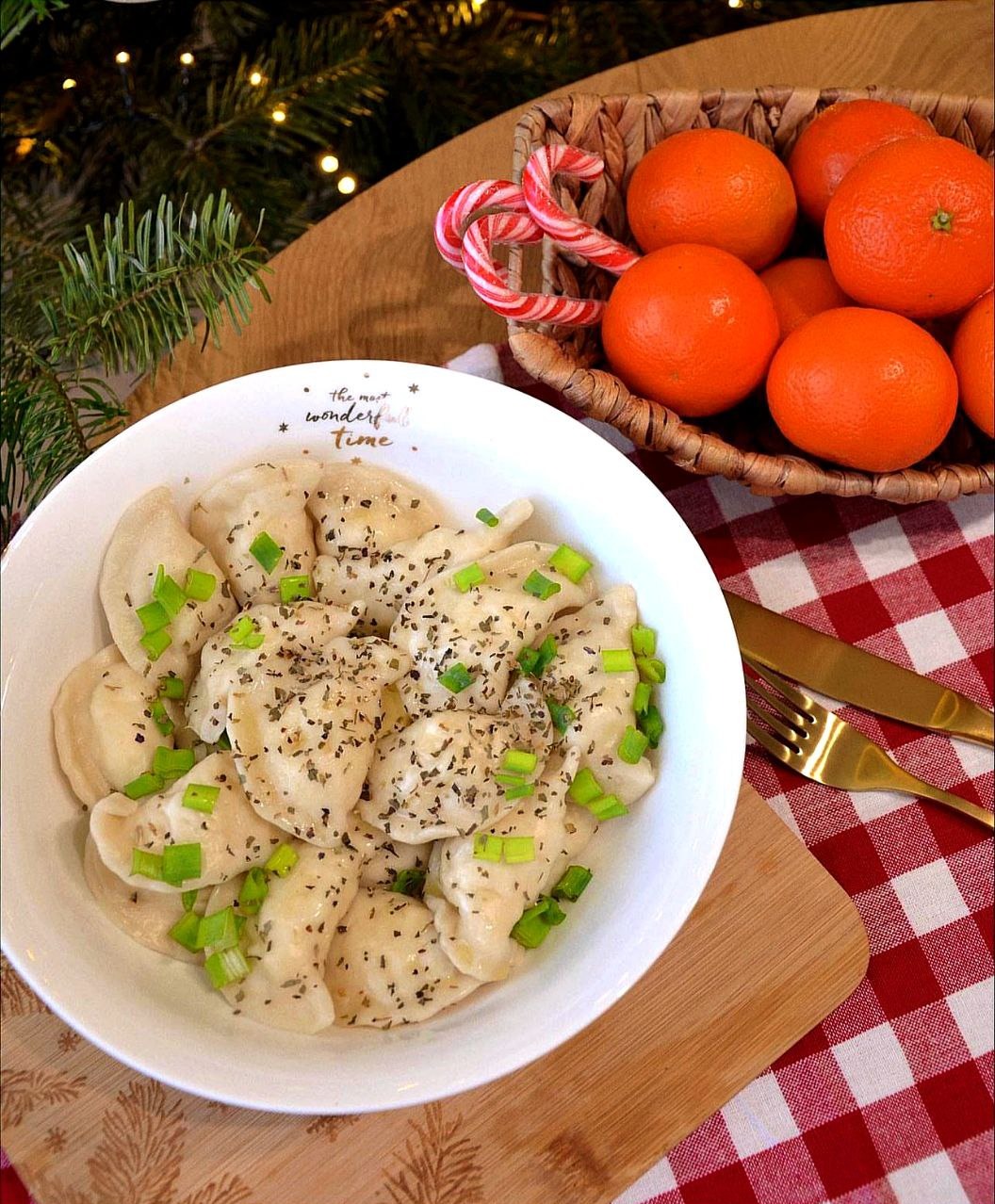 Świąteczny stół z pierogami w białej misce, posypanymi ziołami i zielonym szczypiorkiem, mandarynki w wiklinowym koszyku, laski cukrowe i gałązka choinki w tle.