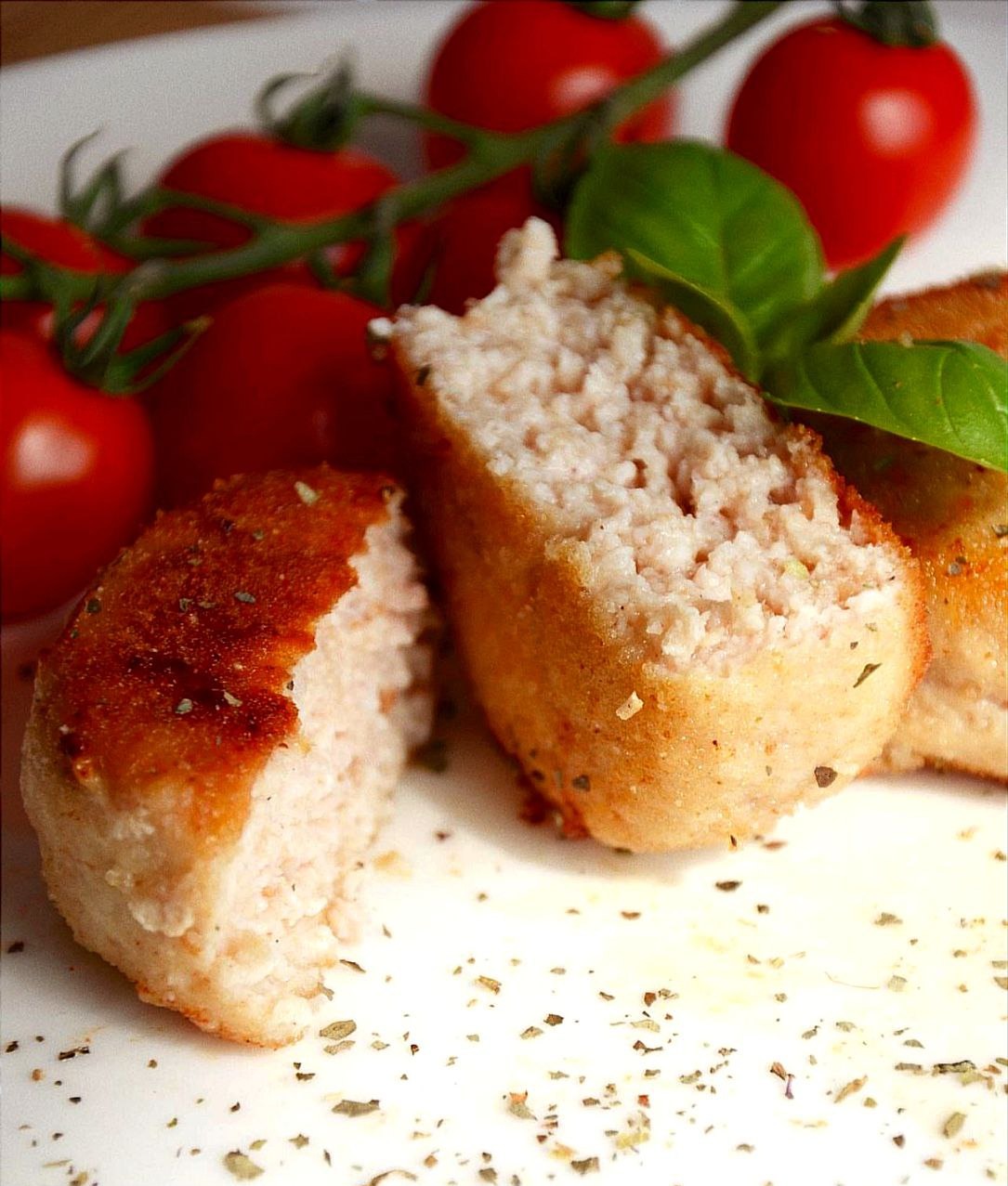 Krokiety z mięsa drobiowego przekrojone na pół, udekorowane gałązką pomidorów cherry i listkiem bazylii, posypane ziołami na białym talerzu.