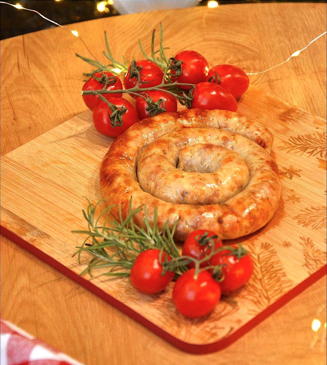 Spiralna, pieczona kiełbasa ułożona na drewnianej desce z motywem świątecznym, udekorowana gałązkami rozmarynu i pomidorkami koktajlowymi na gałązce, oświetlona ciepłym światłem.
