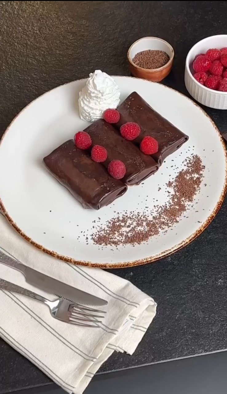 Elegancko podane naleśniki czekoladowe z malinami i bitą śmietaną na białym talerzu z rustykalnym brzegiem, obok sztućce na lnianej serwetce.