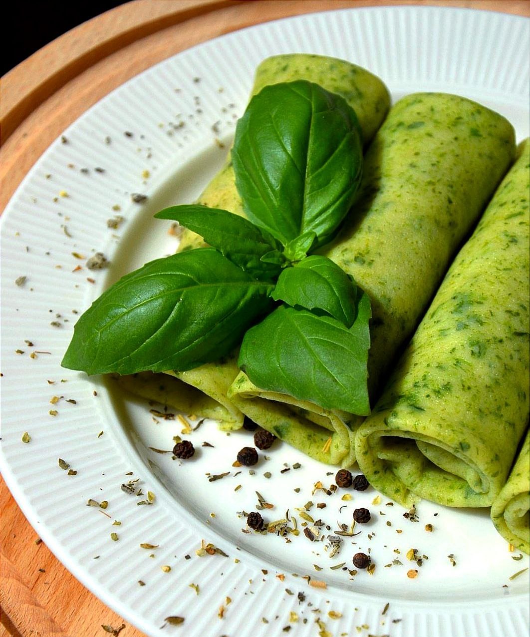 Zielone naleśniki nadziewane, udekorowane świeżą bazylią i przyprawami na białym, karbowanym talerzu, całość na drewnianej desce.