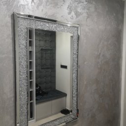 Robert Kalbarczyk - Ściana pokryta dekoracyjną, srebrzystą farbą strukturalną, odbijająca się w lustrze z ramą wysadzaną kryształkami, w tle widoczna szara, tapicerowana wnęka z półkami.