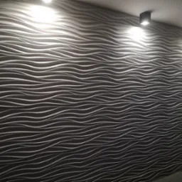 Ściana pokryta dekoracyjnymi panelami 3D w odcieniach szarości, o falistym wzorze, oświetlona przez trzy punktowe lampy sufitowe. W prawym dolnym rogu widoczna roślina doniczkowa.