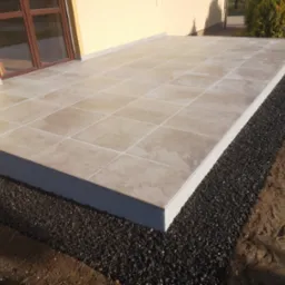 Wykończony taras z beżowych płytek ceramicznych, obrzeże z ciemnego grysu, widok na elewację budynku z brązowymi oknami.