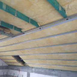 Poddasze w trakcie ocieplania wełną mineralną, widoczne zielone belki drewniane i metalowe profile sufitowe, częściowo zakryte materiałem izolacyjnym.