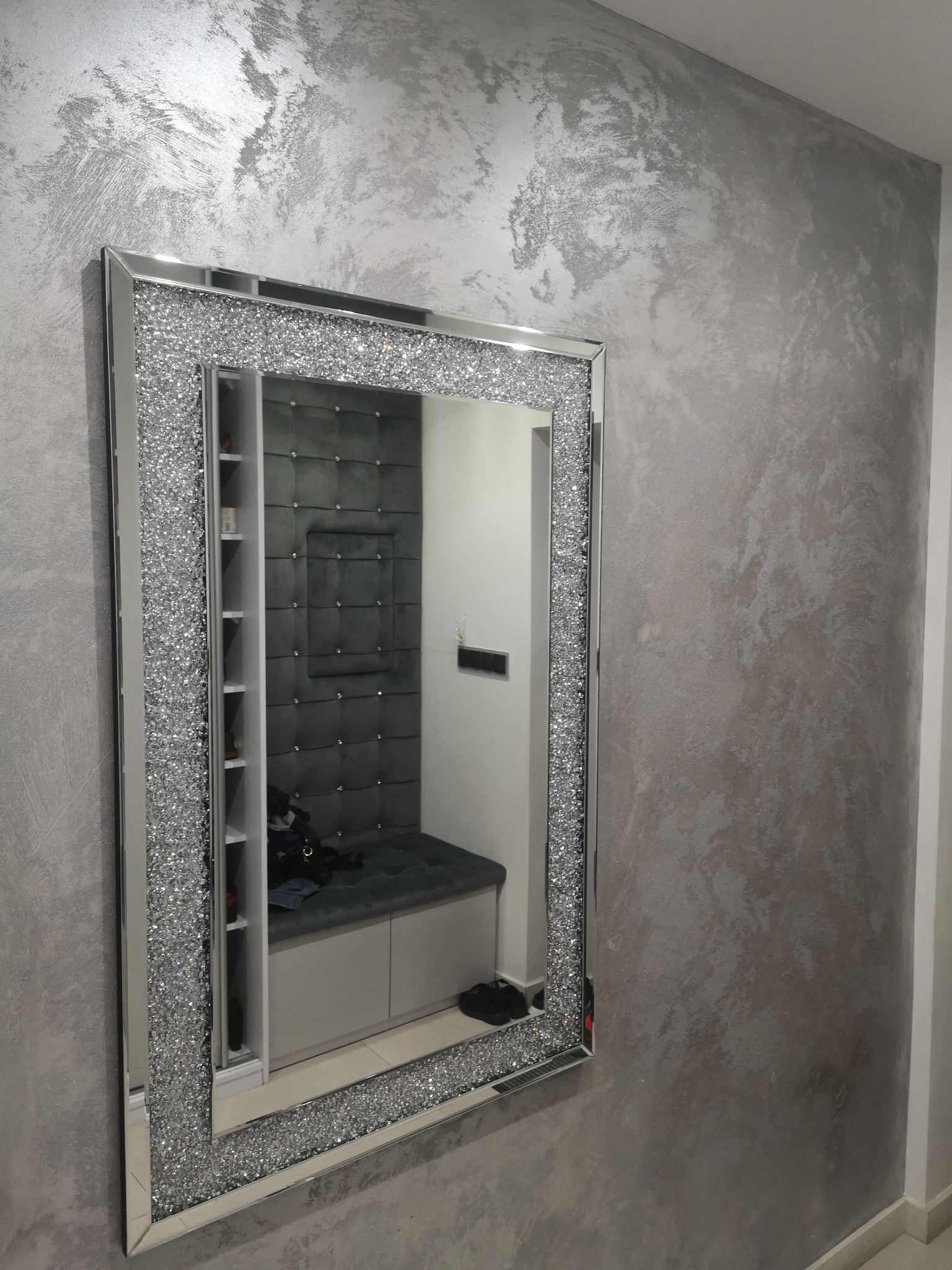 Ściana pokryta dekoracyjną, srebrzystą farbą strukturalną, odbijająca się w lustrze z ramą wysadzaną kryształkami, w tle widoczna szara, tapicerowana wnęka z półkami.