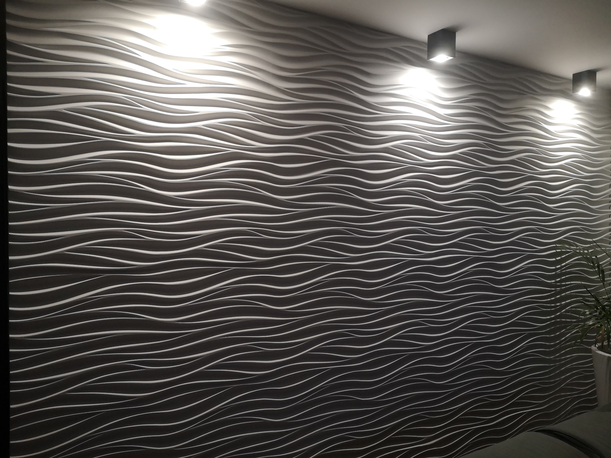 Ściana pokryta dekoracyjnymi panelami 3D w odcieniach szarości, o falistym wzorze, oświetlona przez trzy punktowe lampy sufitowe. W prawym dolnym rogu widoczna roślina doniczkowa.