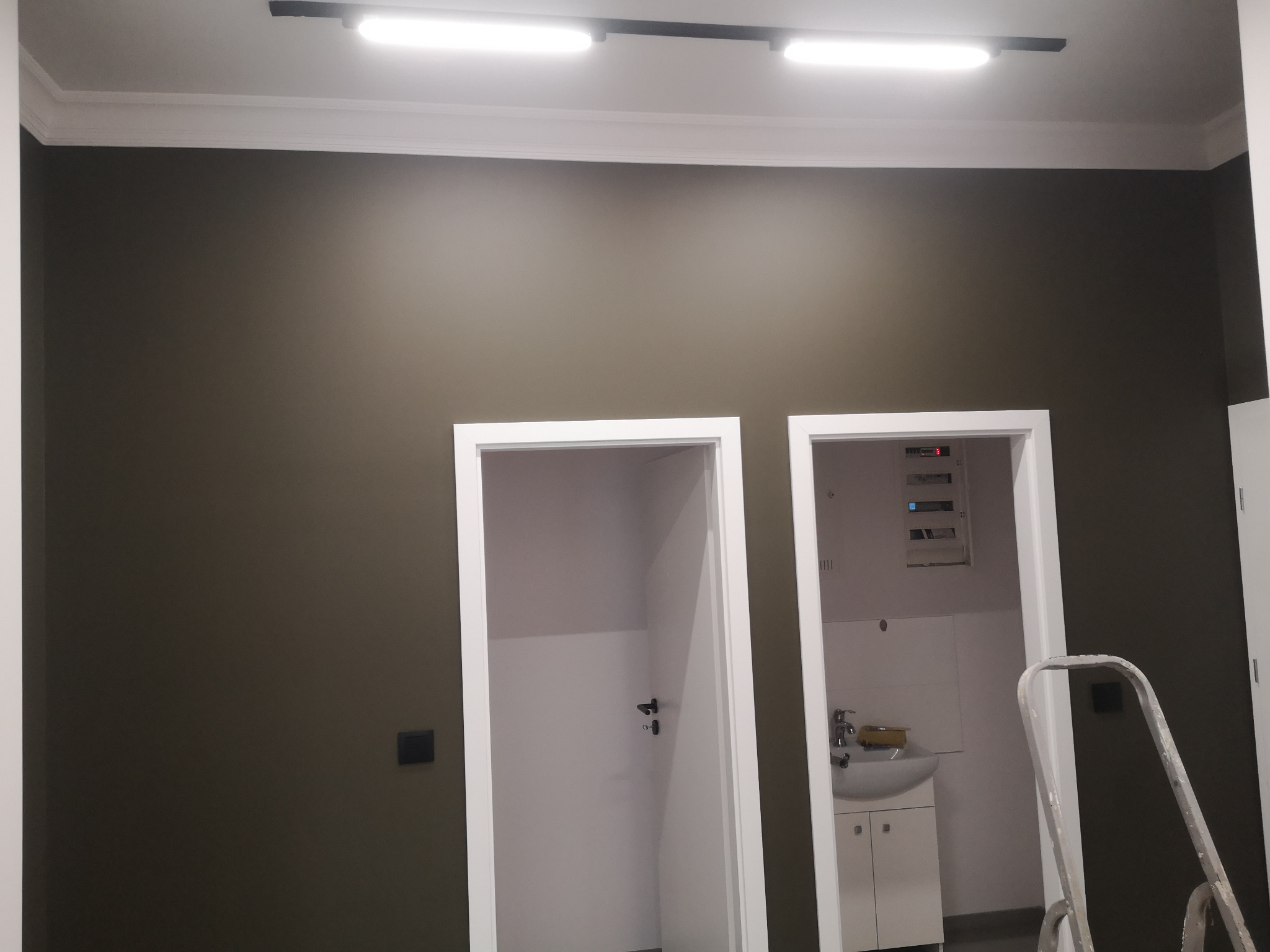 Ściana pomalowana na ciemny kolor w Radomiu, widoczne dwa wejścia do pomieszczeń, białe futryny, listwa dekoracyjna pod sufitem, oświetlenie szynowe, fragment drabiny malarskiej.
