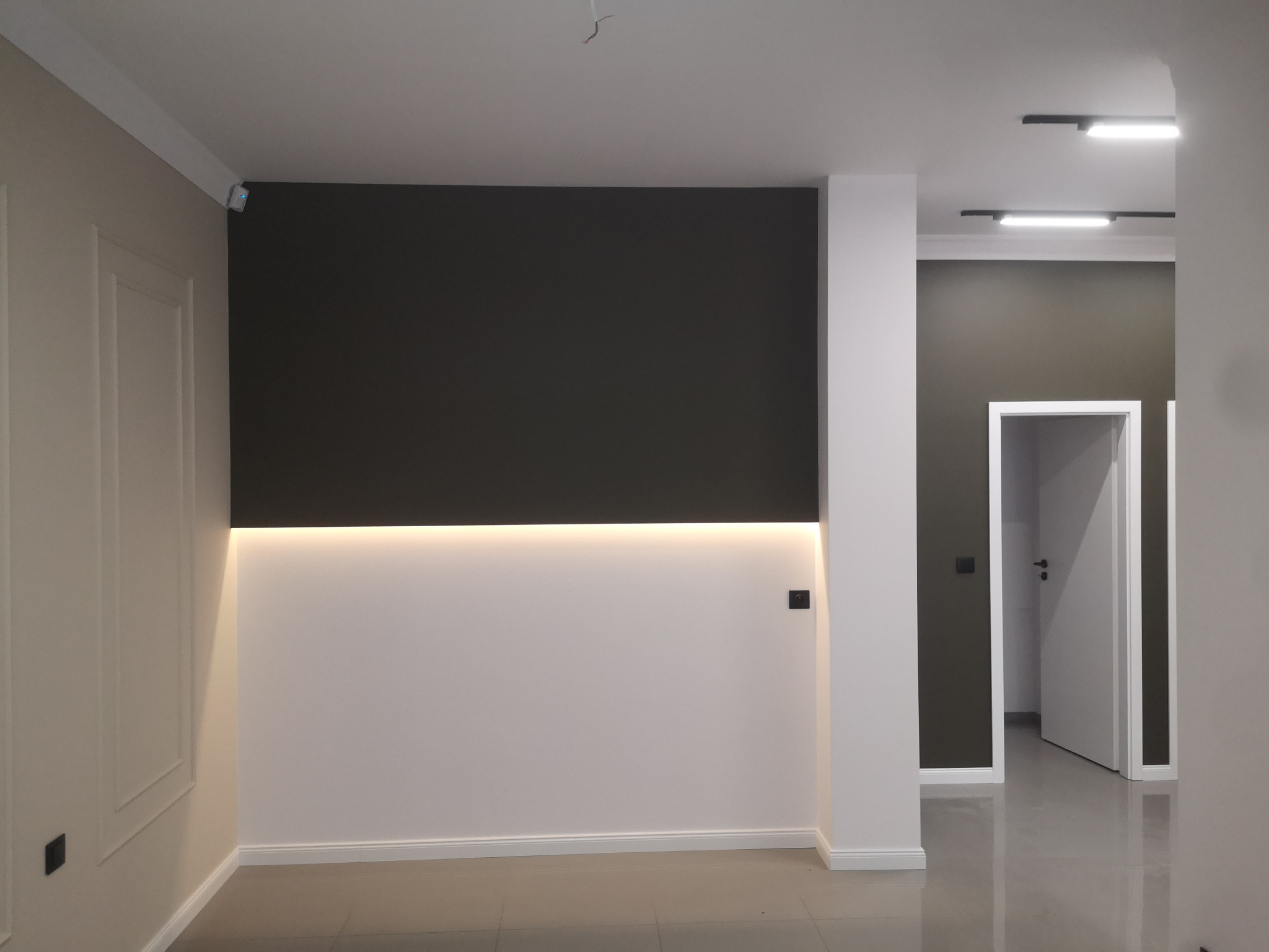 Nowoczesne wnętrze po remoncie: ściana z dwukolorowym malowaniem i podświetleniem LED, widoczne listwy przypodłogowe i gładkie ściany, fragment otwartych drzwi w tle.