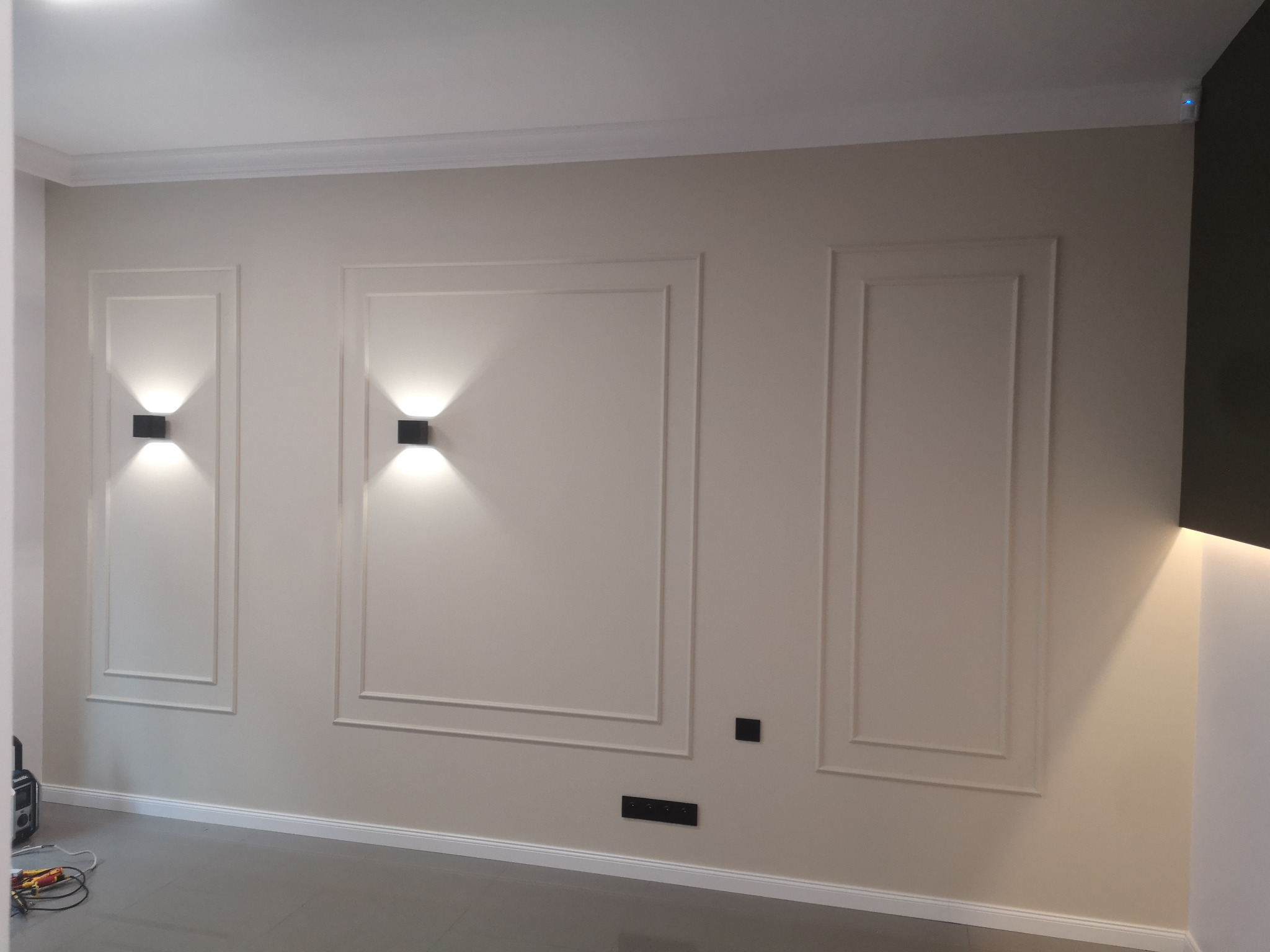 Beżowa ściana z trzema symetrycznymi, dekoracyjnymi ramami, oświetlona czarnymi kinkietami; widoczne gniazdka elektryczne i narzędzia na podłodze.