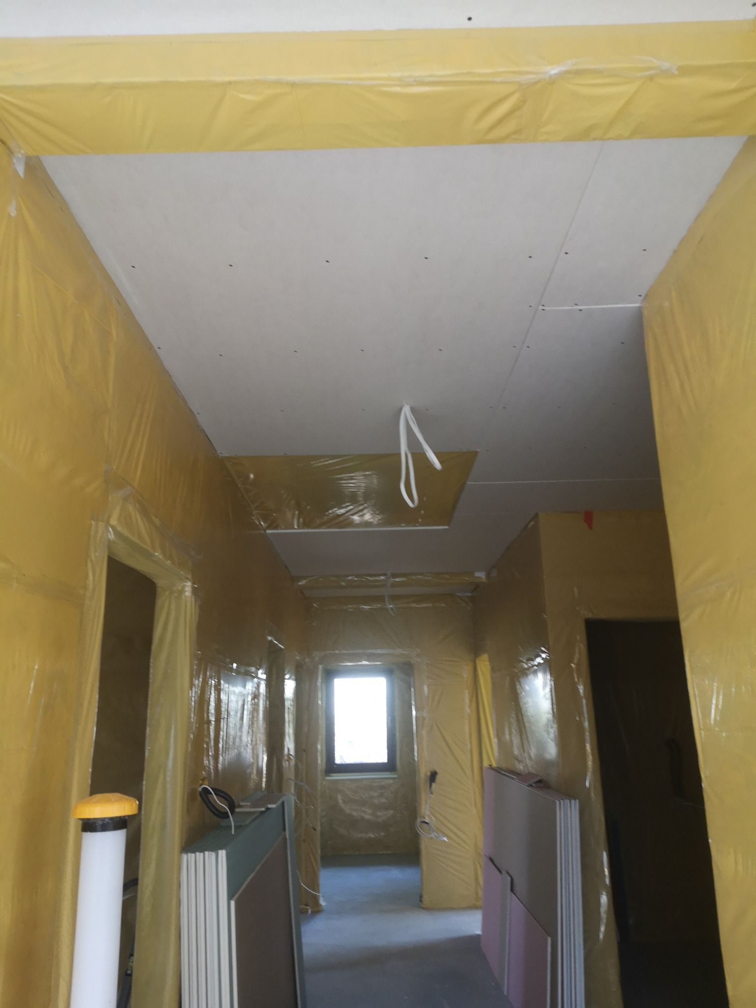 Korytarz w trakcie prac wykończeniowych, ściany zabezpieczone folią, widoczne płyty kartonowo-gipsowe i częściowo zamontowany sufit podwieszany z widocznymi przewodami.