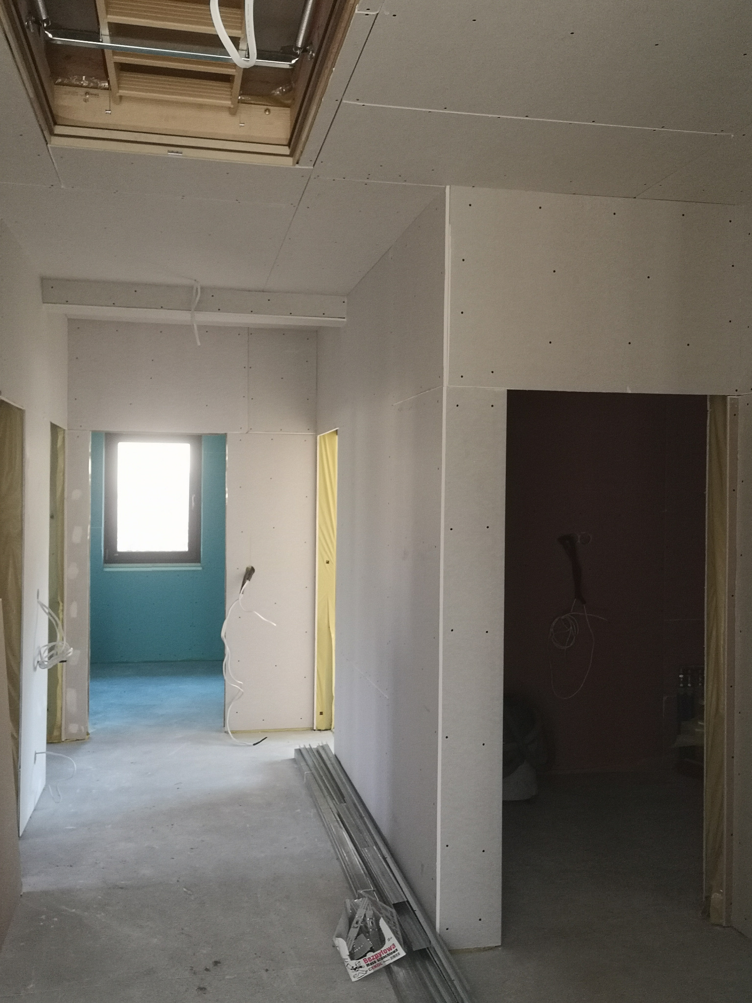 Korytarz w trakcie montażu płyt kartonowo-gipsowych, widoczne profile na podłodze, otwór okienny w suficie oraz instalacja elektryczna.
