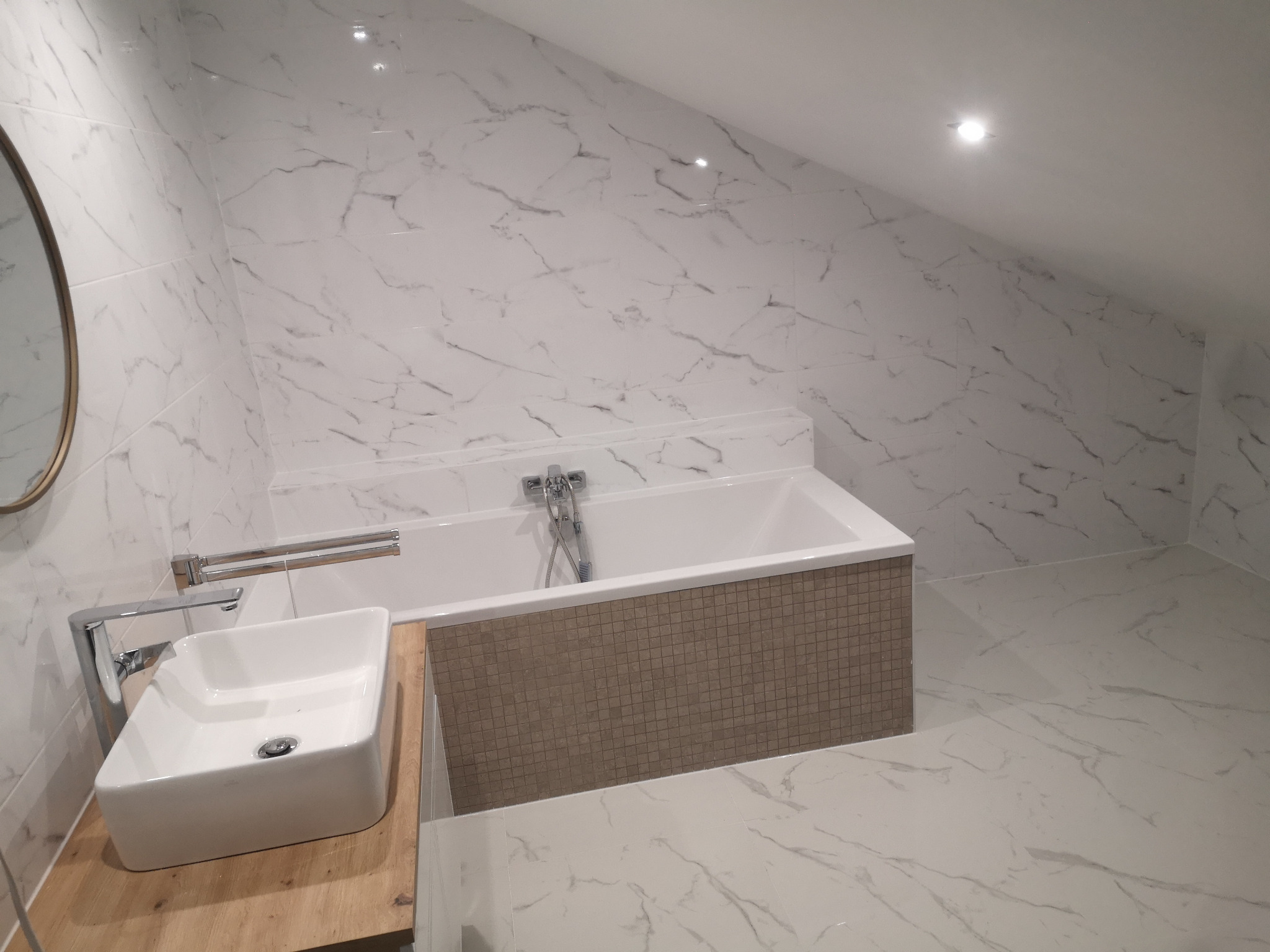 Nowoczesna łazienka na poddaszu z marmurowymi płytkami, białą wanną obudowaną mozaiką, umywalką nablatową na drewnianym blacie i okrągłym lustrem.