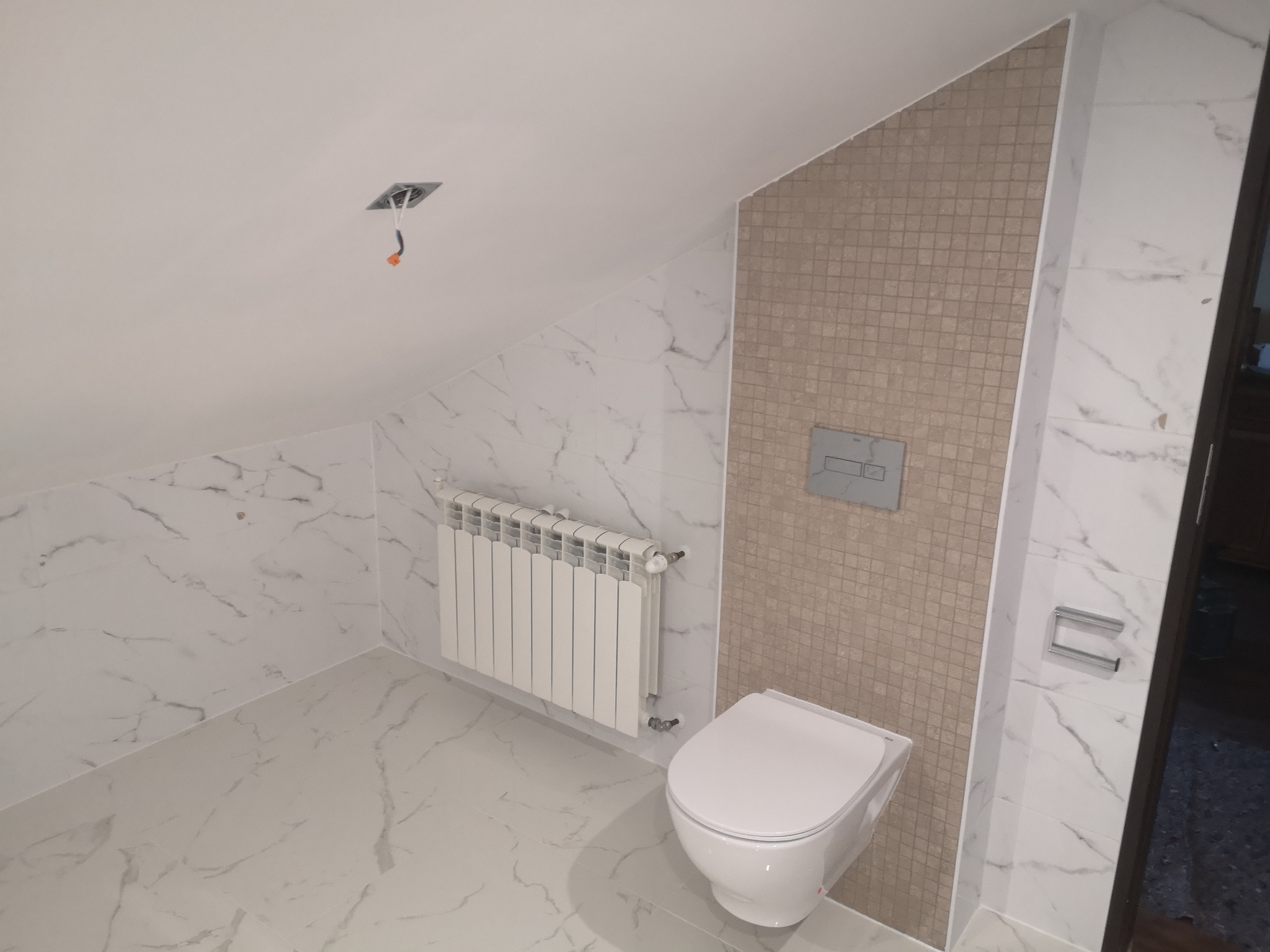 Wykończona łazienka na poddaszu ze skośnym sufitem, białe płytki imitujące marmur na ścianach i podłodze, mozaika w odcieniach beżu na jednej ze ścian, biała toaleta wisząca i grzejnik.