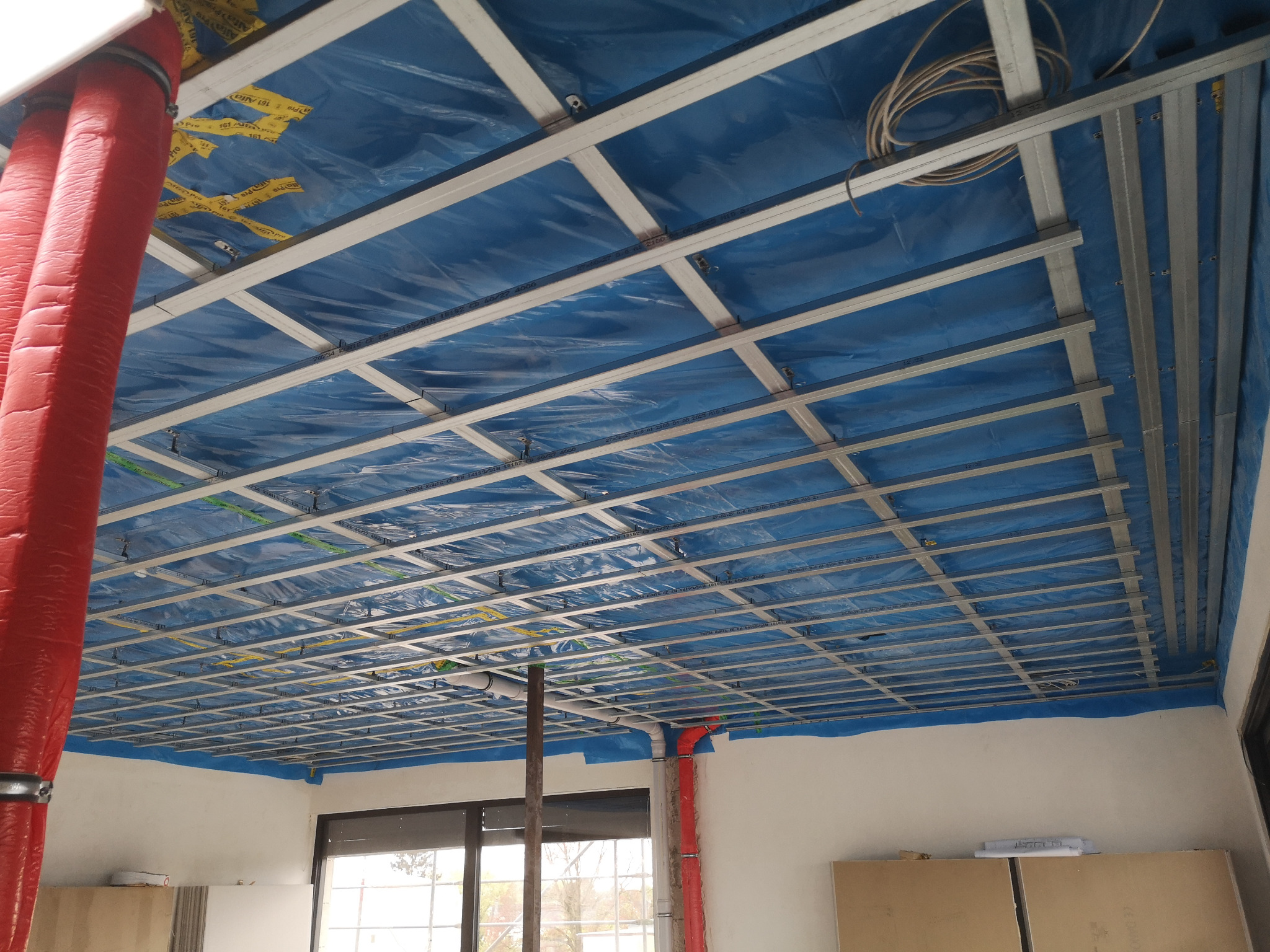 Montaż metalowej konstrukcji sufitu podwieszanego z zabezpieczeniem folią budowlaną, widoczne instalacje elektryczne i rury wentylacyjne w osłonie.