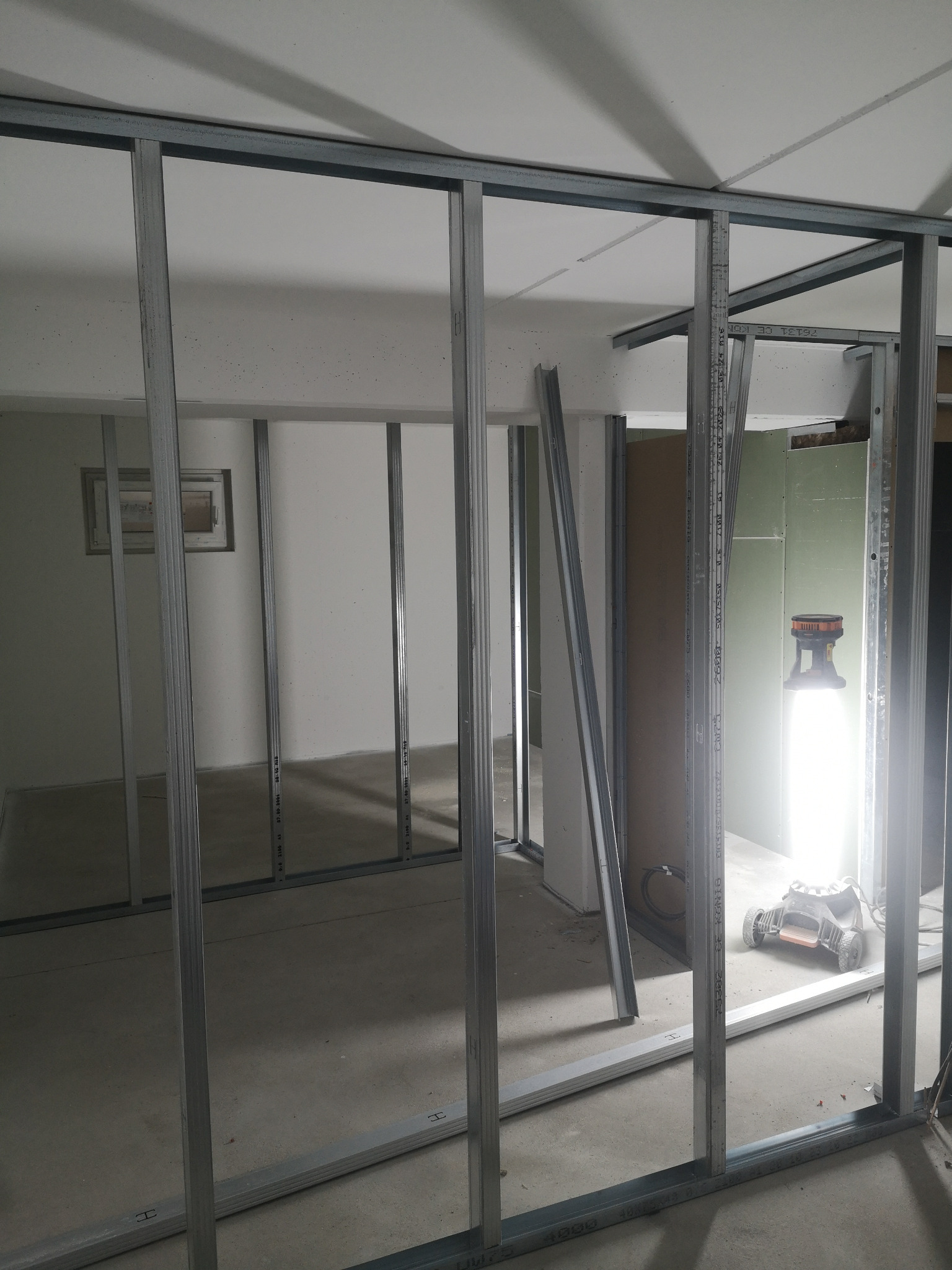 Metalowy szkielet ścian działowych z profili stalowych w trakcie montażu, widoczna lampa budowlana oświetlająca przestrzeń, częściowo obłożona ściana z płyty karton-gips.