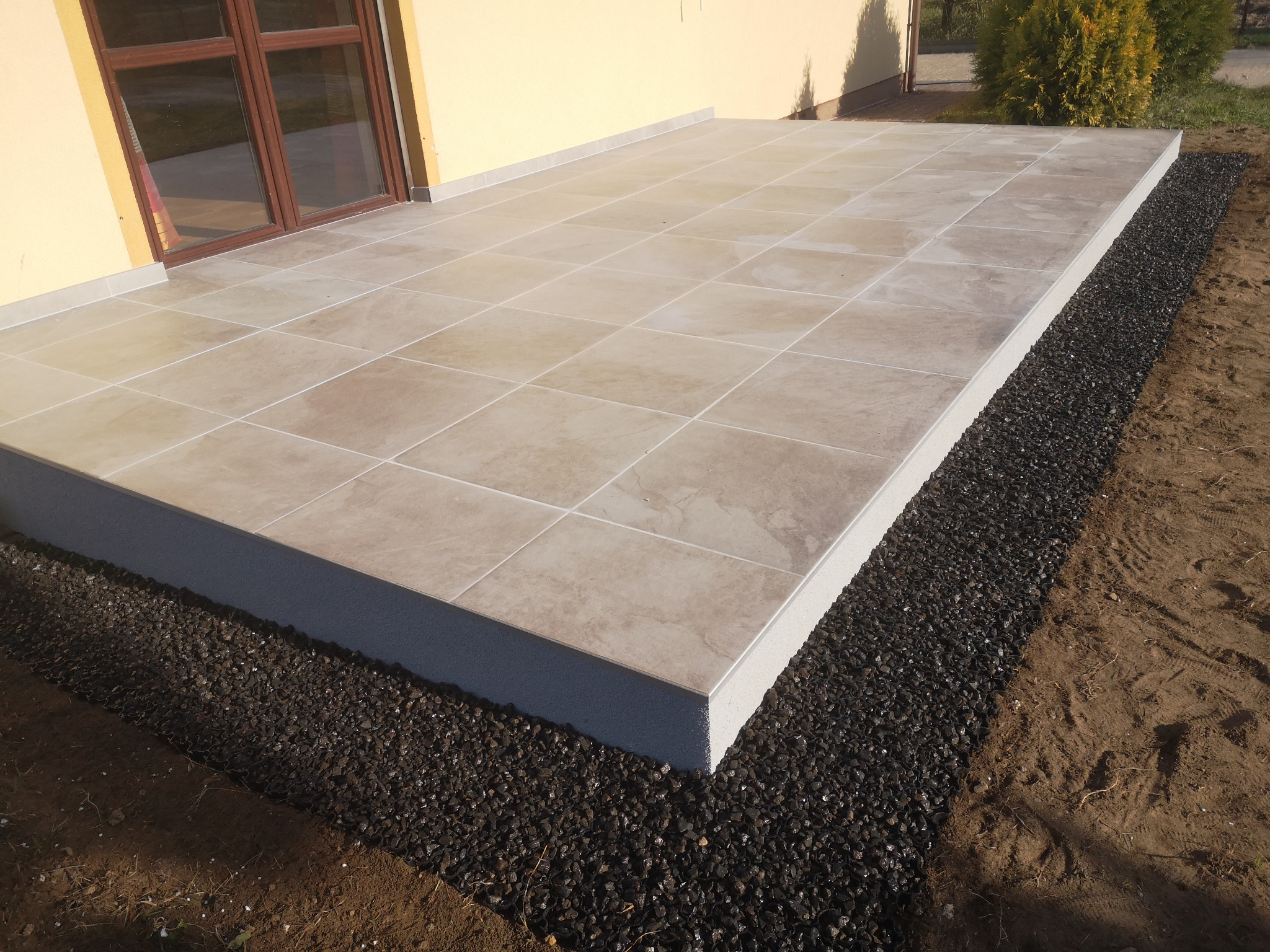 Wykończony taras z beżowych płytek ceramicznych, obrzeże z ciemnego grysu, widok na elewację budynku z brązowymi oknami.