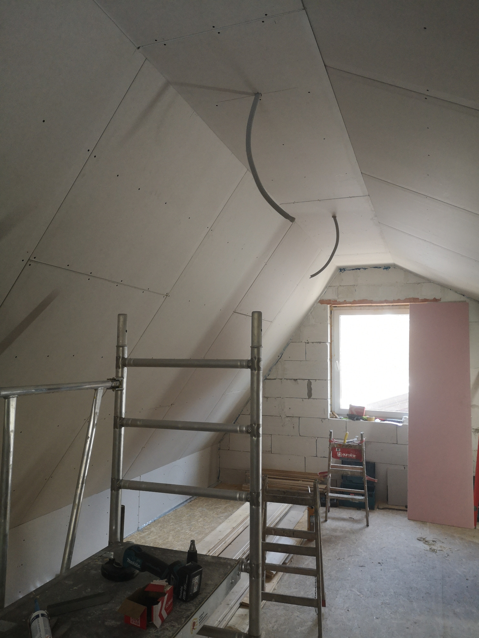 Montaż płyt karton gips na poddaszu, widoczny fragment instalacji elektrycznej, rusztowanie, okno i bloczki betonowe.
