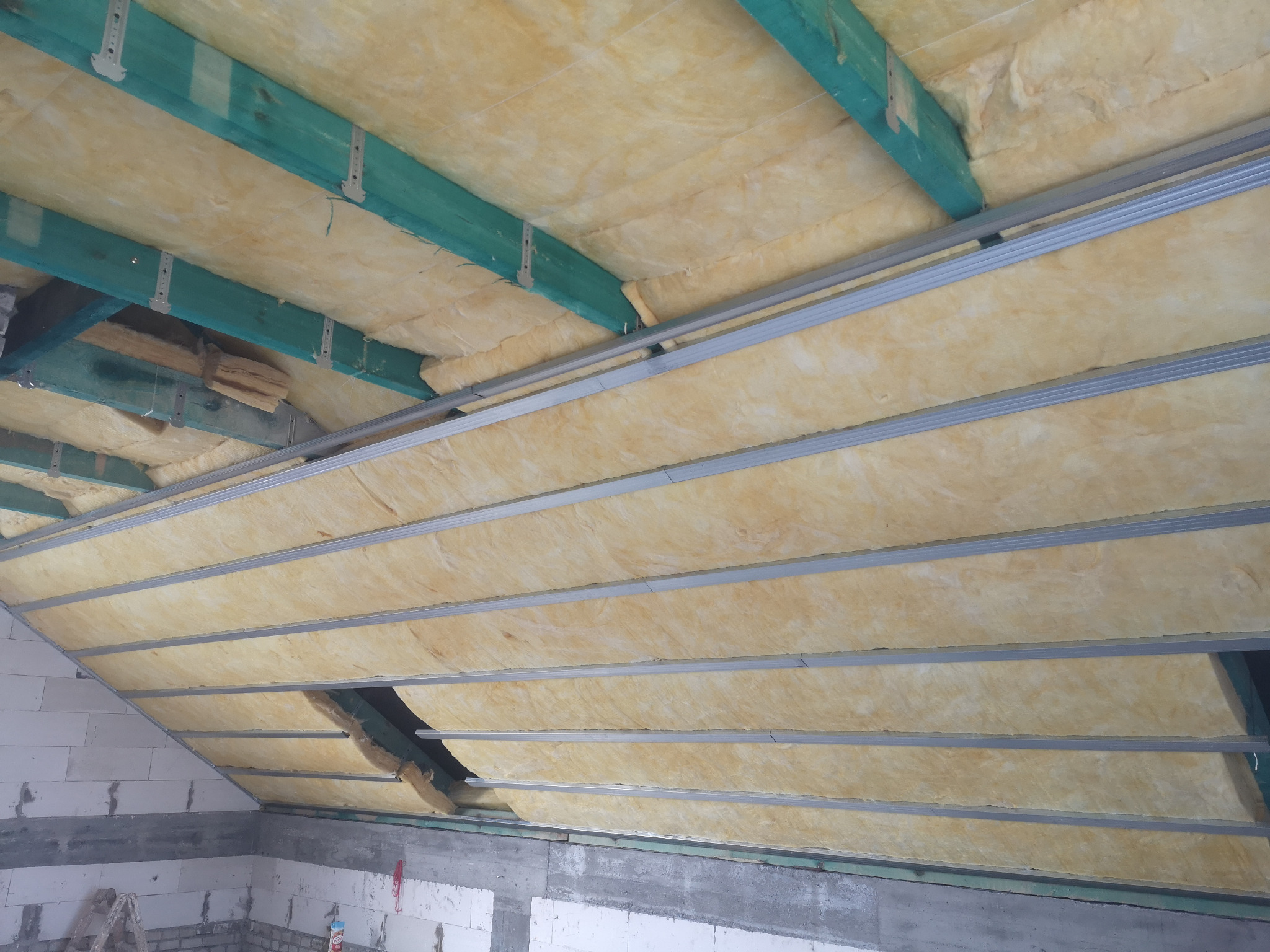 Poddasze w trakcie ocieplania wełną mineralną, widoczne zielone belki drewniane i metalowe profile sufitowe, częściowo zakryte materiałem izolacyjnym.