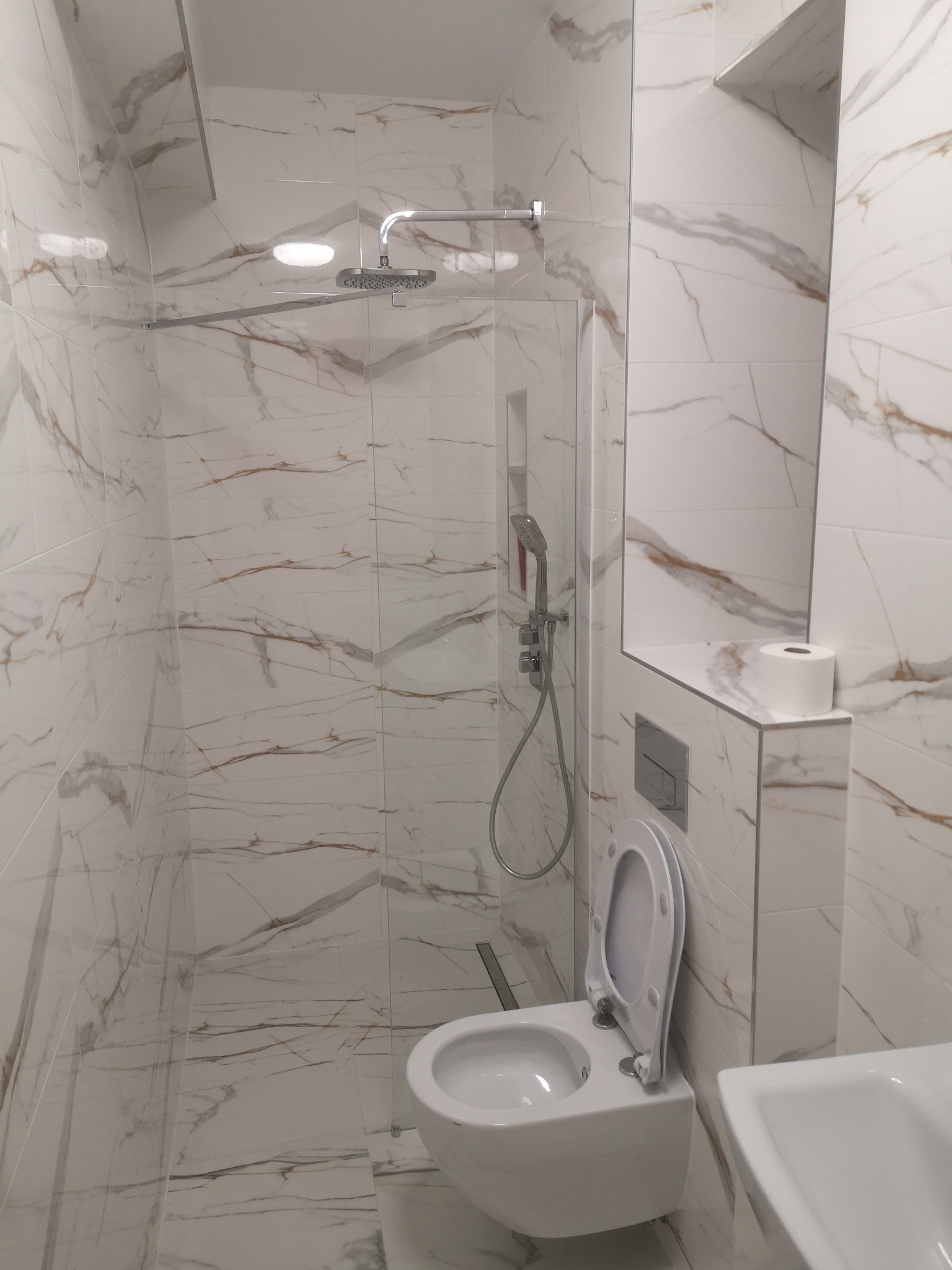 Biała łazienka z kabiną typu walk-in, wykończona marmurowymi płytkami z brązowymi żyłami. Widoczna toaleta podtynkowa i deszczownica.