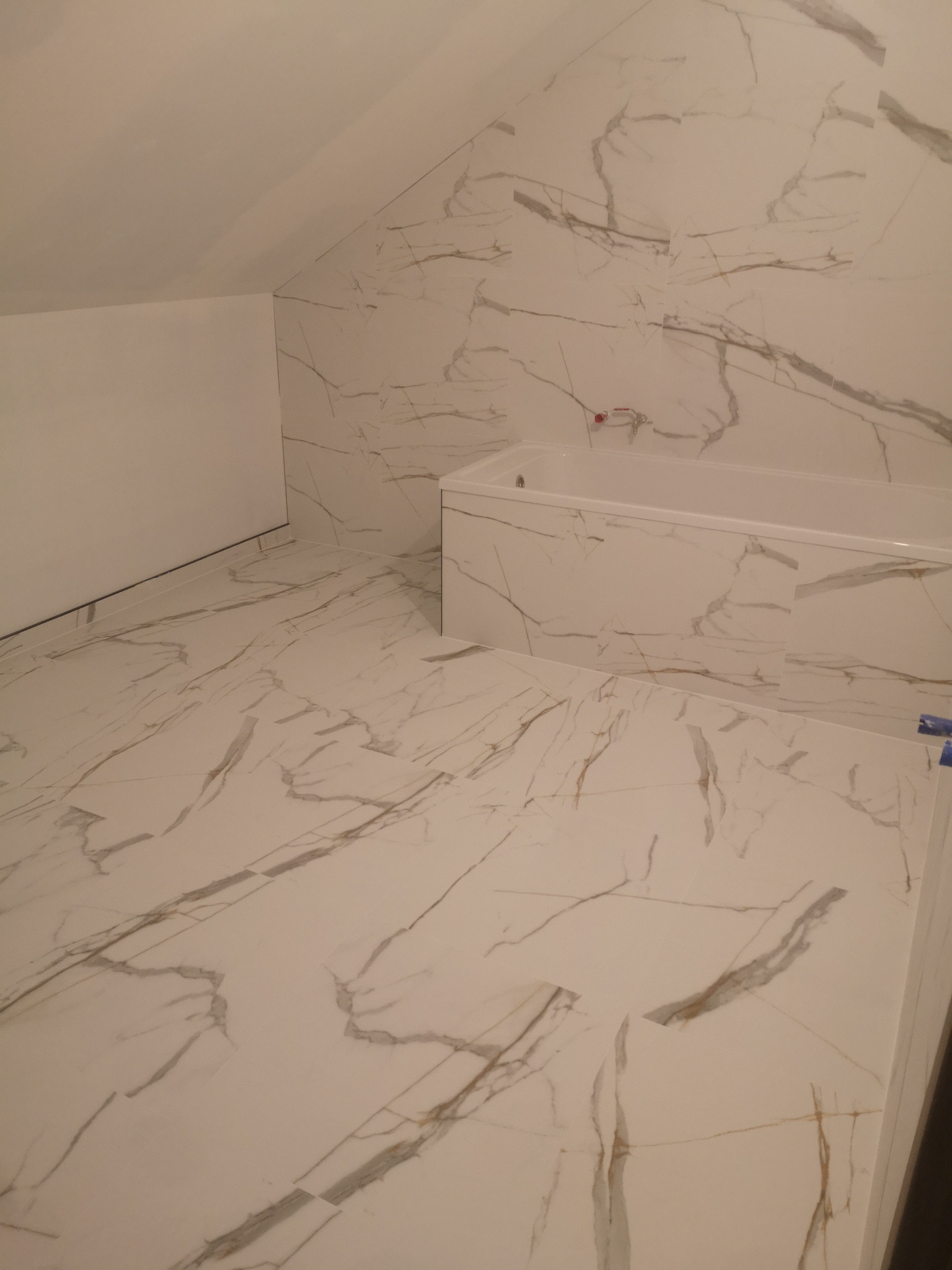 Wykończona łazienka na poddaszu z wanną, wyłożona białymi płytkami imitującymi marmur z szarymi żyłami na podłodze i ścianach. Widoczne skosy dachu.