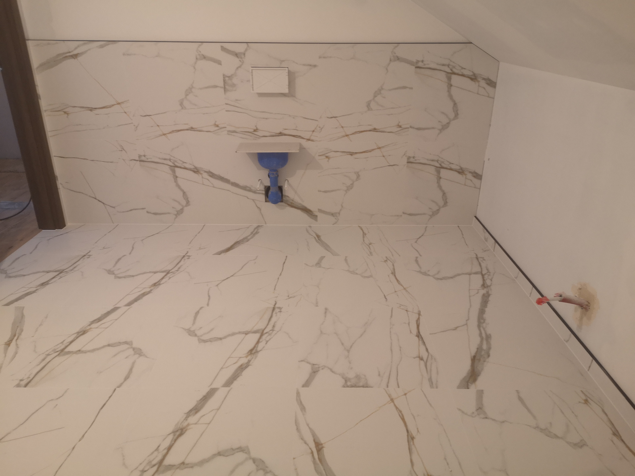 Wykończona łazienka na poddaszu z białymi płytkami imitującymi marmur na ścianach i podłodze. Widoczna instalacja podtynkowa WC i wystająca rurka.