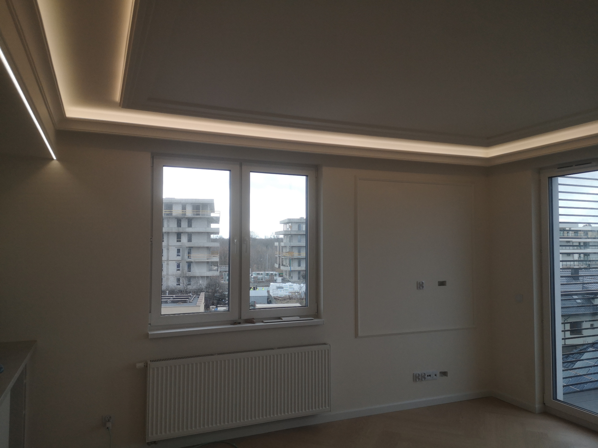 Jasne wnętrze po remoncie: okno z widokiem na budowę, grzejnik, listwy LED na suficie i ścianach, gładkie ściany. Minimalistyczny design z elementami dekoracyjnymi.