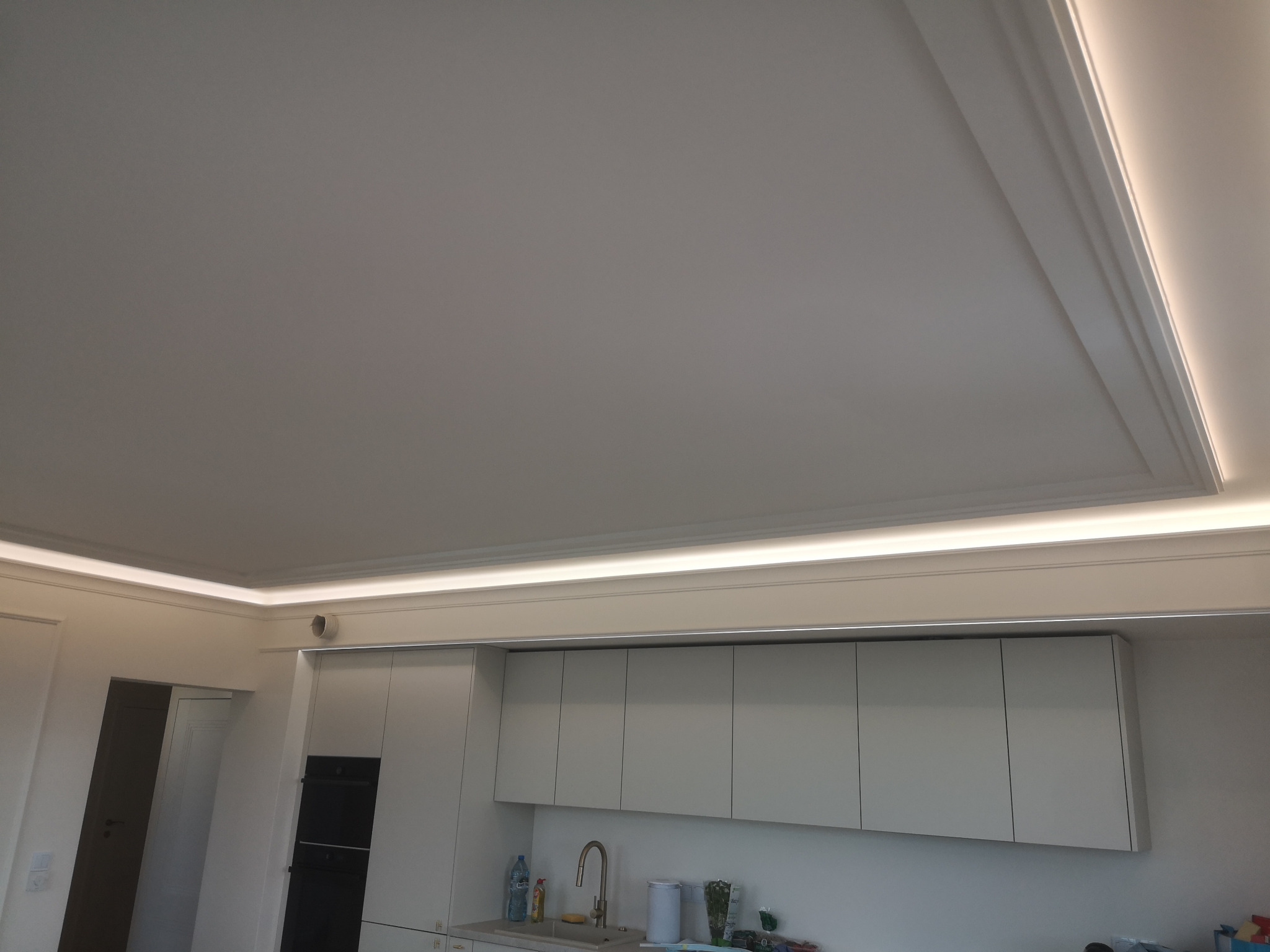 Biały sufit z dekoracyjną listwą i oświetleniem LED w nowoczesnej kuchni. Minimalistyczne szafki wiszące w tle. Ujęcie z dołu, podkreślające detale wykończenia.