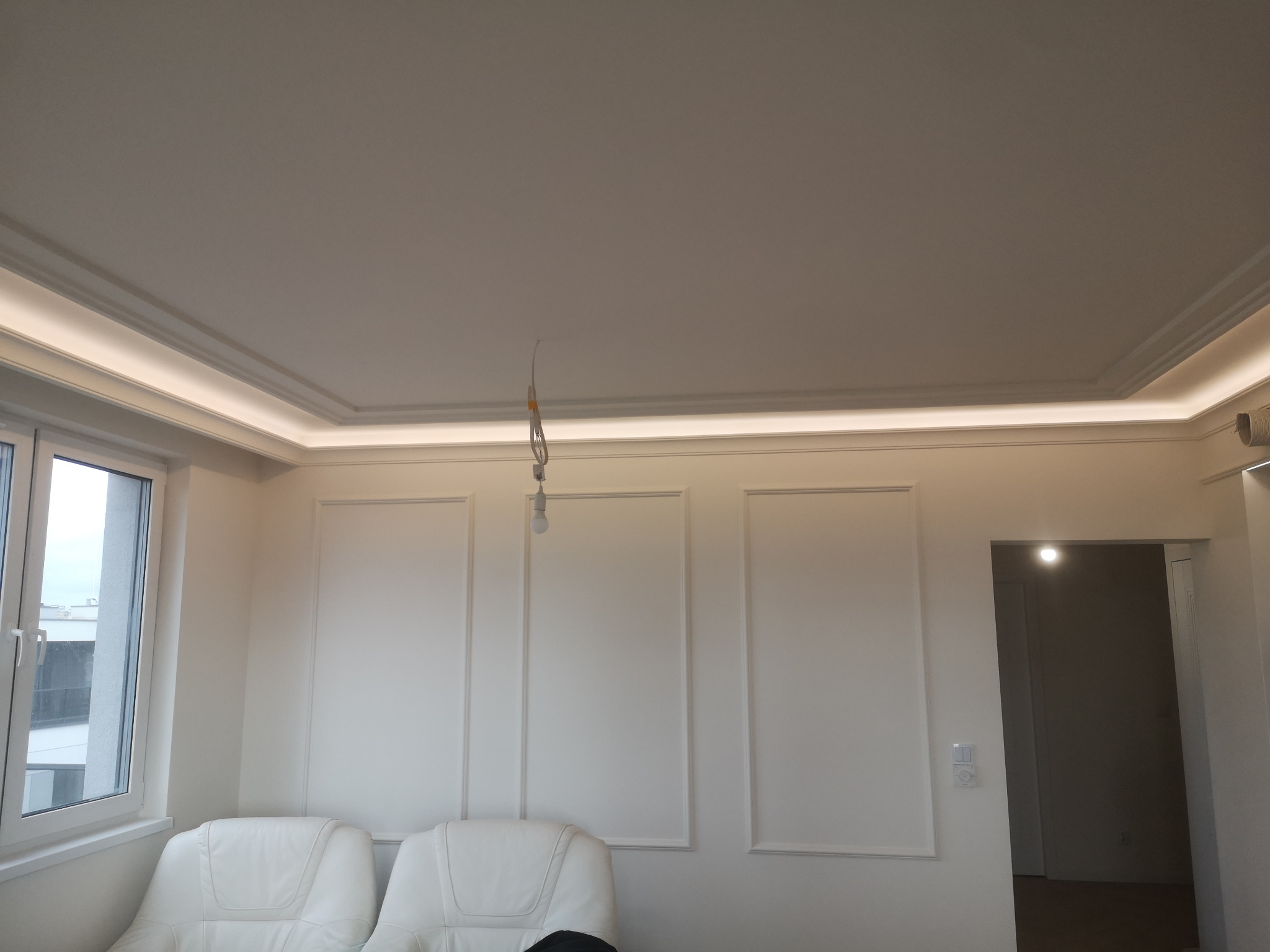 Białe wnętrze po wykończeniu: sufit z oświetleniem LED, ściany z dekoracyjnymi panelami, okno i dwa białe fotele. Widoczne niedokończone podłączenie lampy.