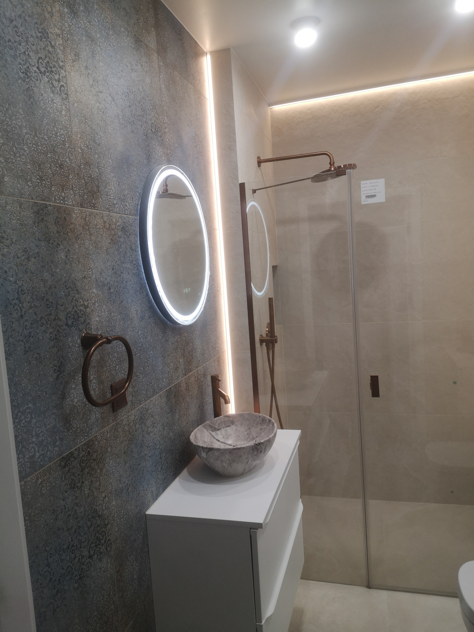 Nowoczesna łazienka z szarymi płytkami o wzorzystej strukturze, okrągłym lustrem z podświetleniem LED i kabiną prysznicową ze złotymi elementami.