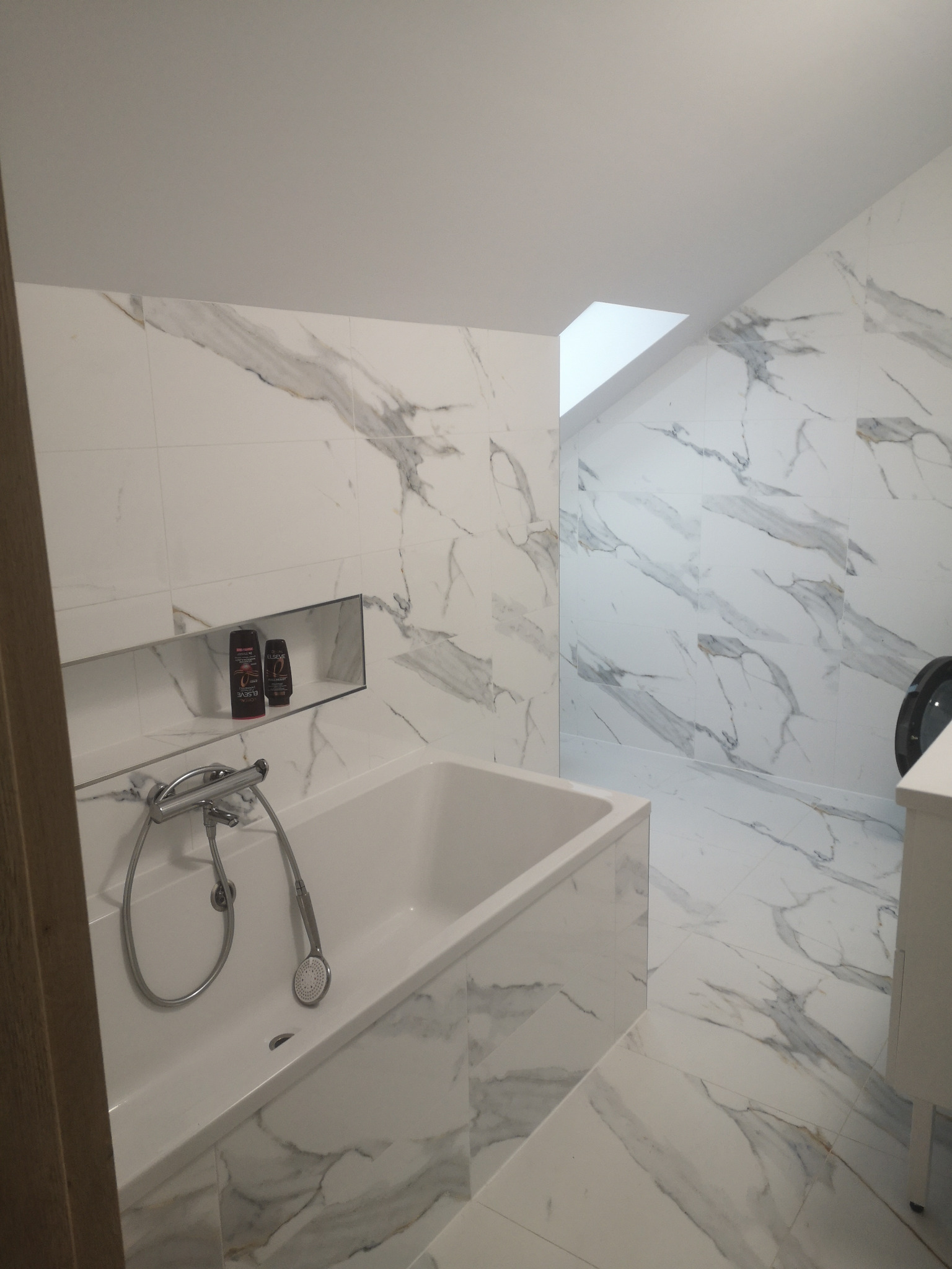 Biała łazienka na poddaszu z wanną, armaturą i marmurowymi płytkami. W ścianie wnęka z kosmetykami. Ukośny sufit z oknem dachowym. Nowoczesny design.