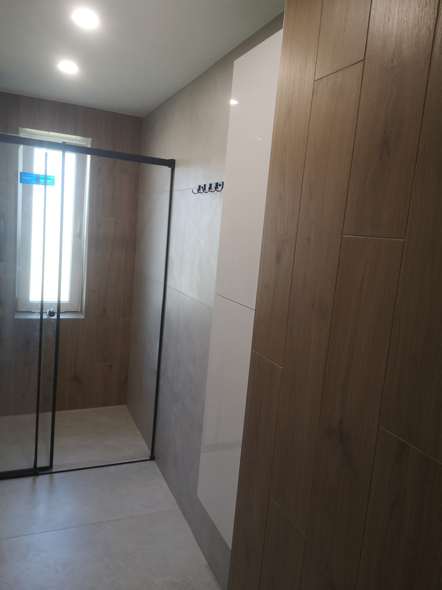 Nowoczesna łazienka z kabiną prysznicową typu walk-in, wykończona płytkami imitującymi drewno i jasnymi panelami. Minimalistyczne wieszaki na ręczniki.