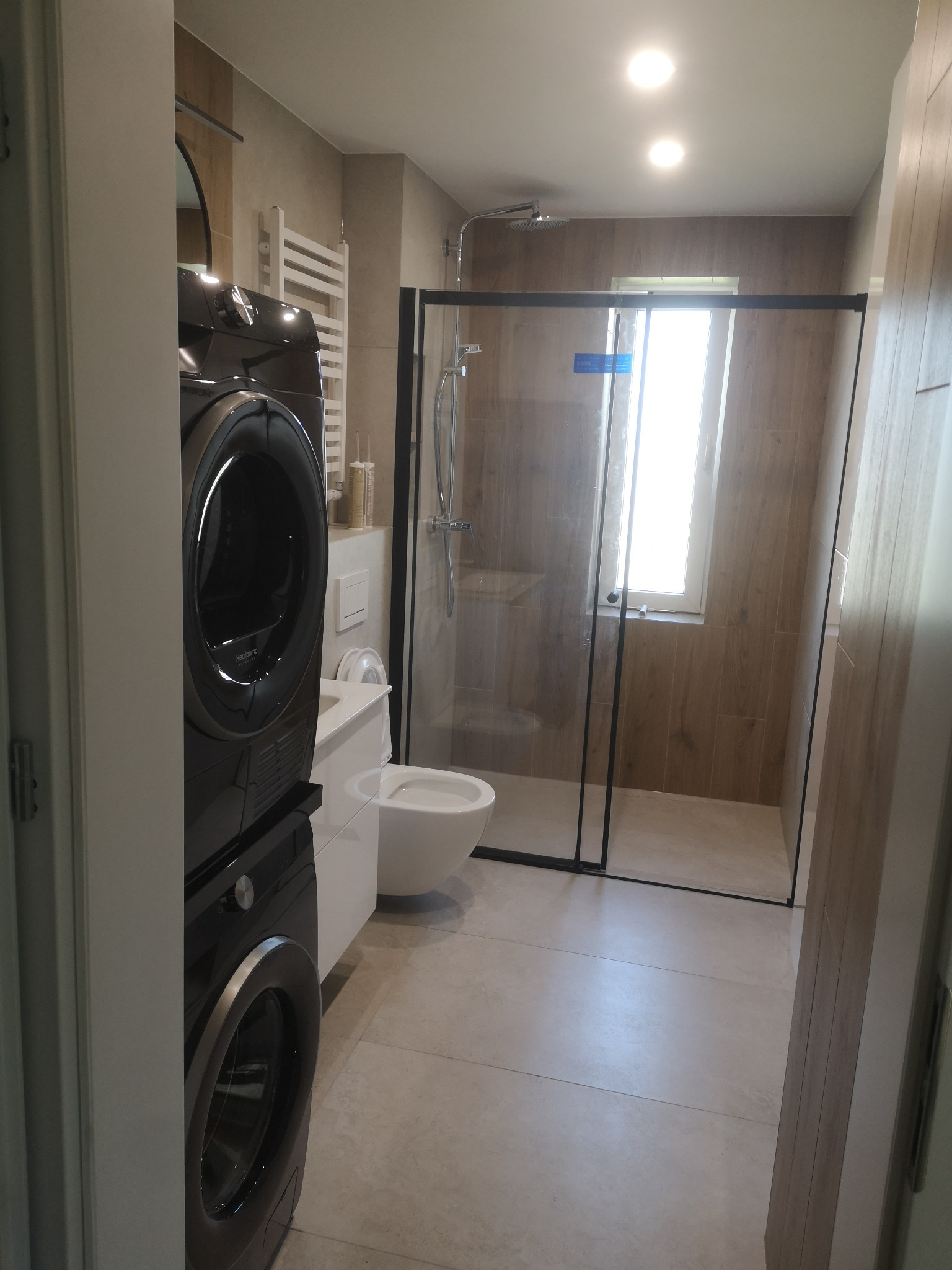Nowoczesna łazienka z kabiną prysznicową typu walk-in, podwieszaną toaletą i pralko-suszarką w kolorze czarnym. Płytki imitujące drewno na ścianach i szare płytki podłogowe.
