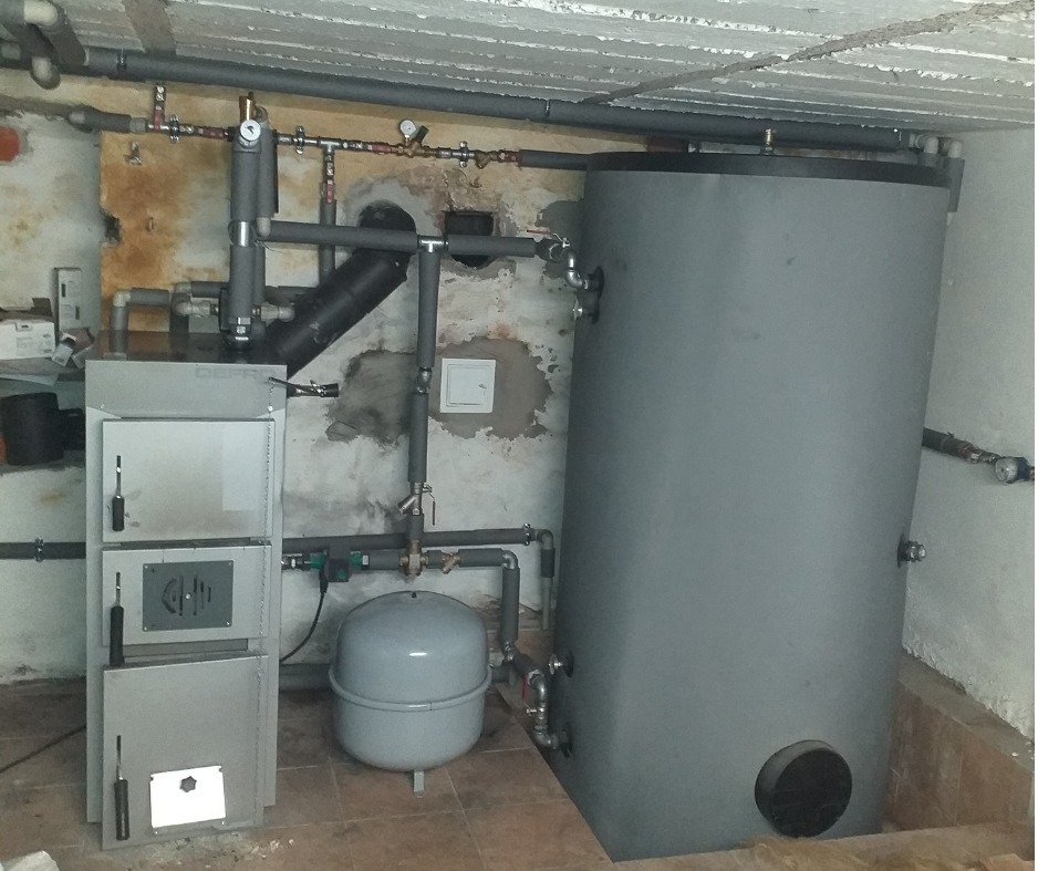 Instalacja grzewcza w piwnicy: piec na paliwo stałe, zbiornik akumulacyjny i naczynie wzbiorcze połączone rurami. Widoczne ślady wilgoci na ścianach.