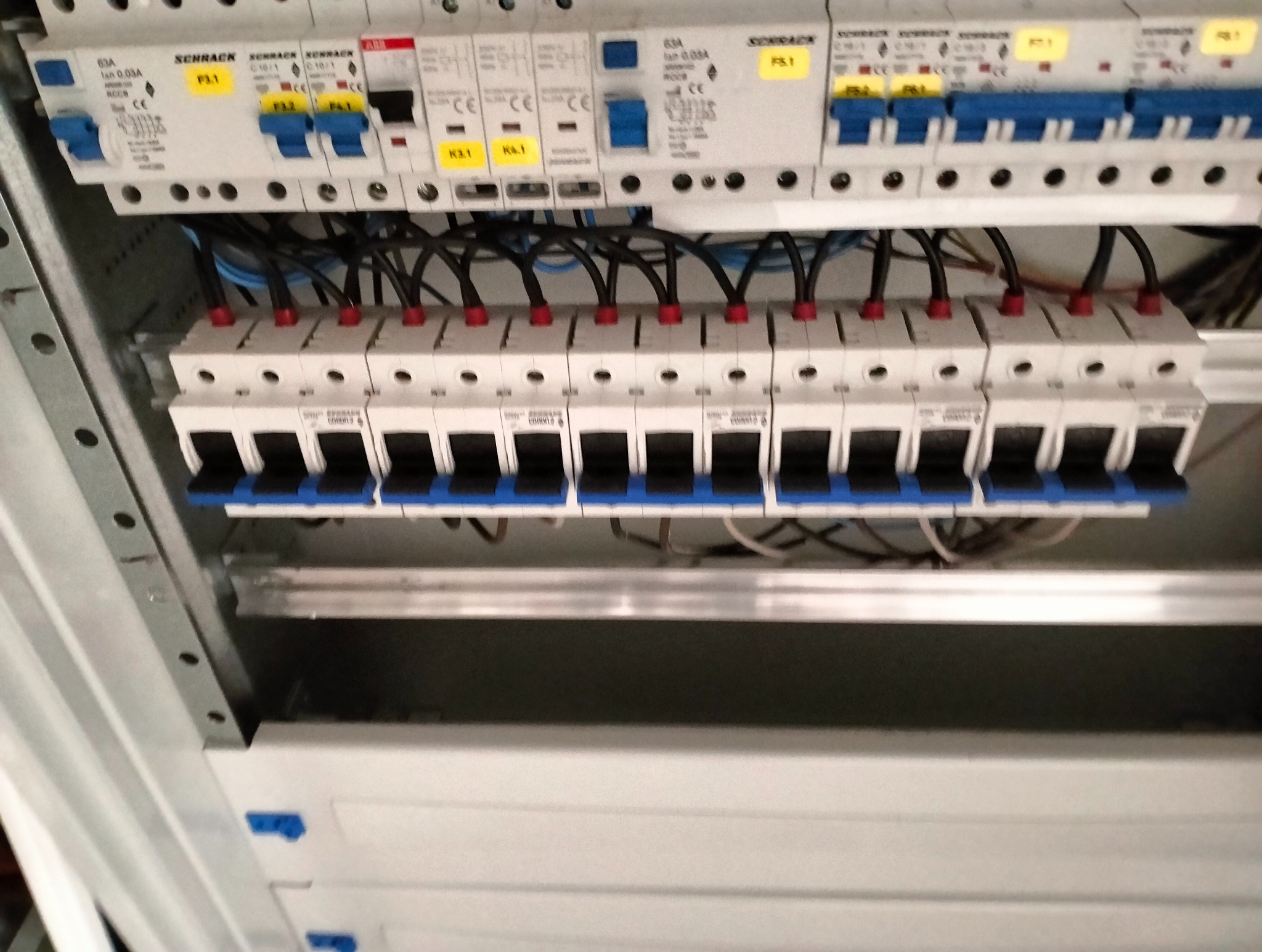 Rozdzielnia elektryczna z widocznymi bezpiecznikami i przewodami wewnątrz metalowej obudowy. Górny rząd bezpieczników marki Schrack i ABB z żółtymi etykietami.