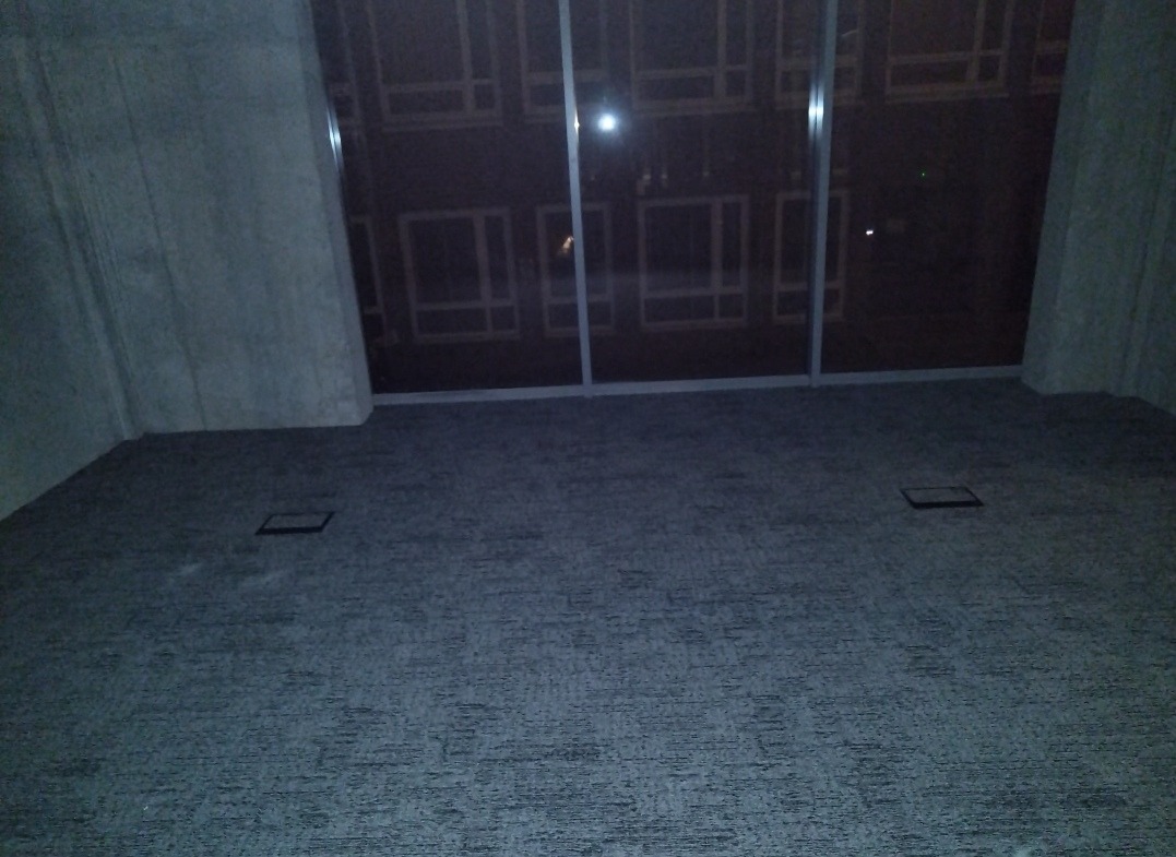 Pokój z szarą wykładziną, widok na okno z budynkami w tle. Dwa otwory w podłodze sugerują instalacje elektryczne. Nocne oświetlenie.