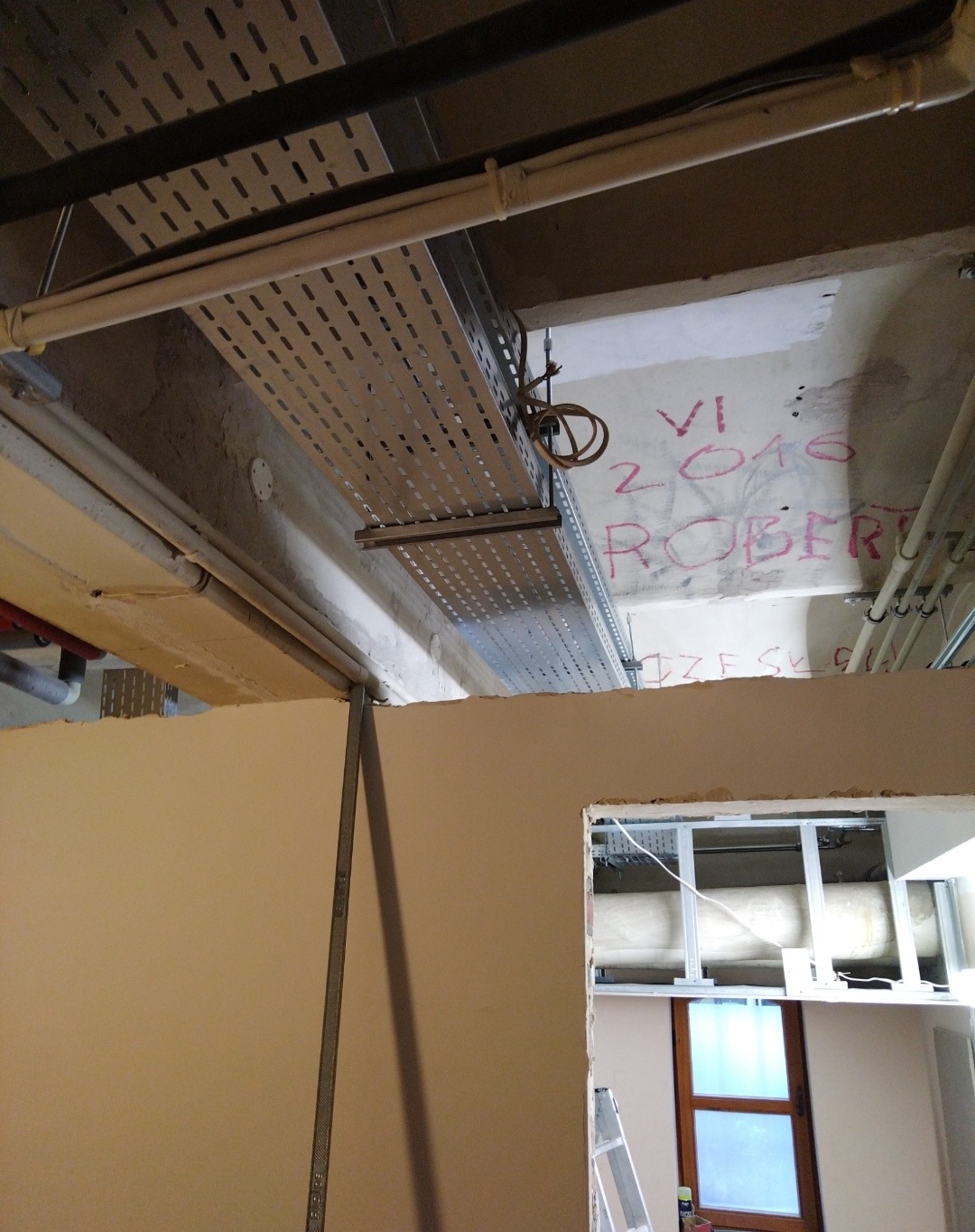 Metalowa korytka kablowa pod sufitem w trakcie prac remontowych. Widoczne rury, ściany w surowym stanie i fragment okna. Na suficie napisy.