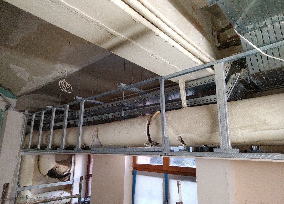 Montaż podwieszanego sufitu z widocznymi rurami wentylacyjnymi i metalowymi profilami, w trakcie prac wykończeniowych. Widoczne okna i przewody elektryczne.