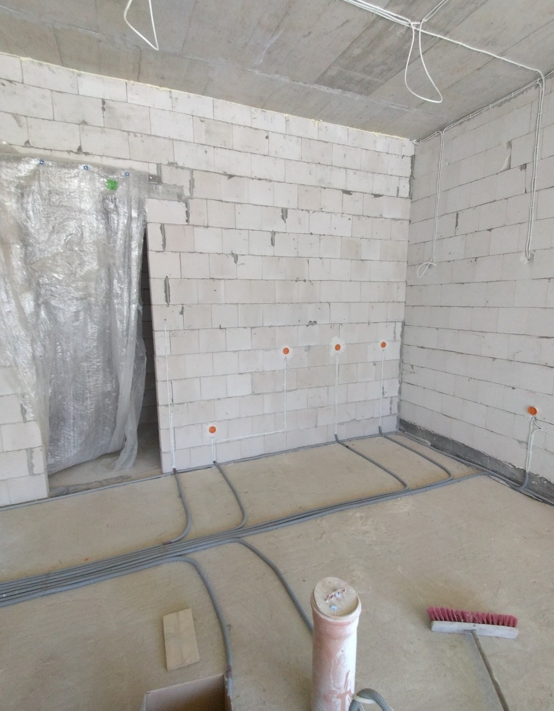 Surowy stan: instalacja elektryczna w nowym budynku. Szare rury na podłodze, kable w ścianach z bloczków, zabezpieczone folią drzwi.