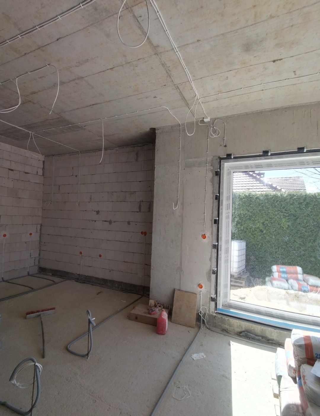 Surowy stan wnętrza z widoczną instalacją elektryczną na suficie i ścianach. W tle okno z widokiem na zieleń. Na podłodze materiały budowlane.