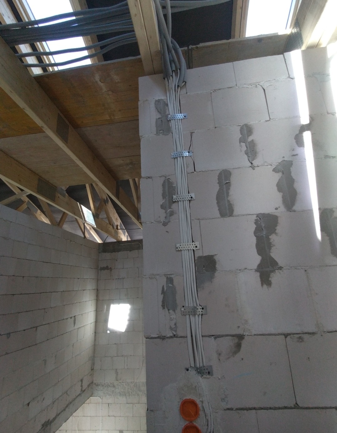 Instalacja elektryczna w surowym budynku: kable w peszlach mocowane do ściany z betonu komórkowego, widoczne puszki instalacyjne i drewniana konstrukcja dachu.