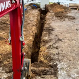 Czerwona minikoparka Yanmar częściowo widoczna obok wąskiego wykopu w ziemi z resztkami śniegu, w tle budynek i drzewa.