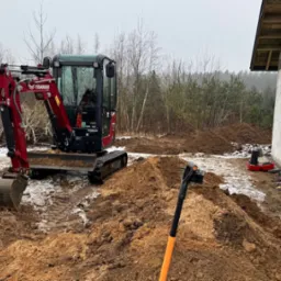 Czerwona minikoparka Yanmar na placu budowy, obok fundamentów domu z bloczków, z widoczną izolacją przeciwwilgociową i resztkami śniegu na ziemi, z łopatą wbita w pryzmę ziemi na pierwszym planie.