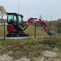 EŁKOP - Czerwono-czarna minikoparka Yanmar 2.2 stoi na trawiastym terenie za metalowym ogrodzeniem, w tle widoczna droga ekspresowa i skrzynka elektryczna. Pochmurny dzień.