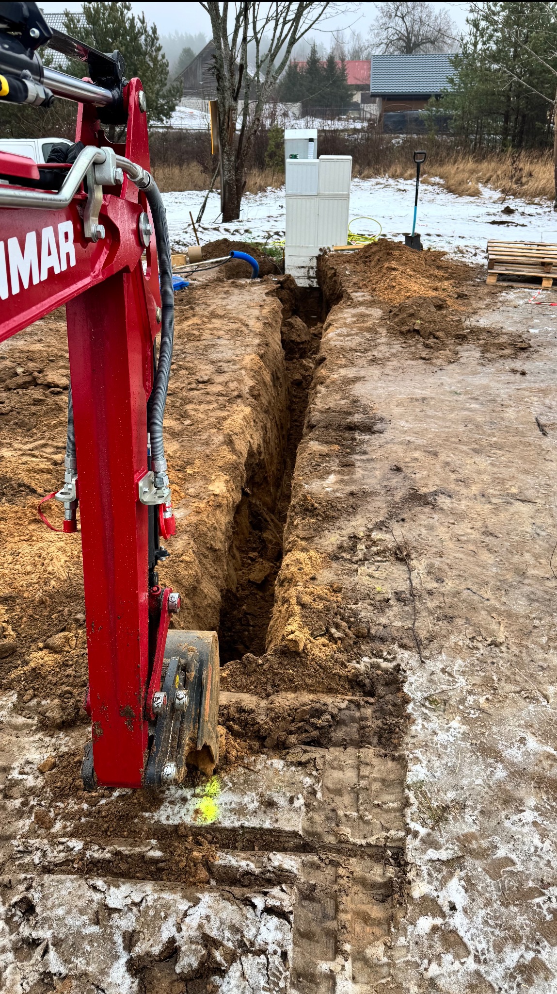 Czerwona minikoparka Yanmar częściowo widoczna obok wąskiego wykopu w ziemi z resztkami śniegu, w tle budynek i drzewa.
