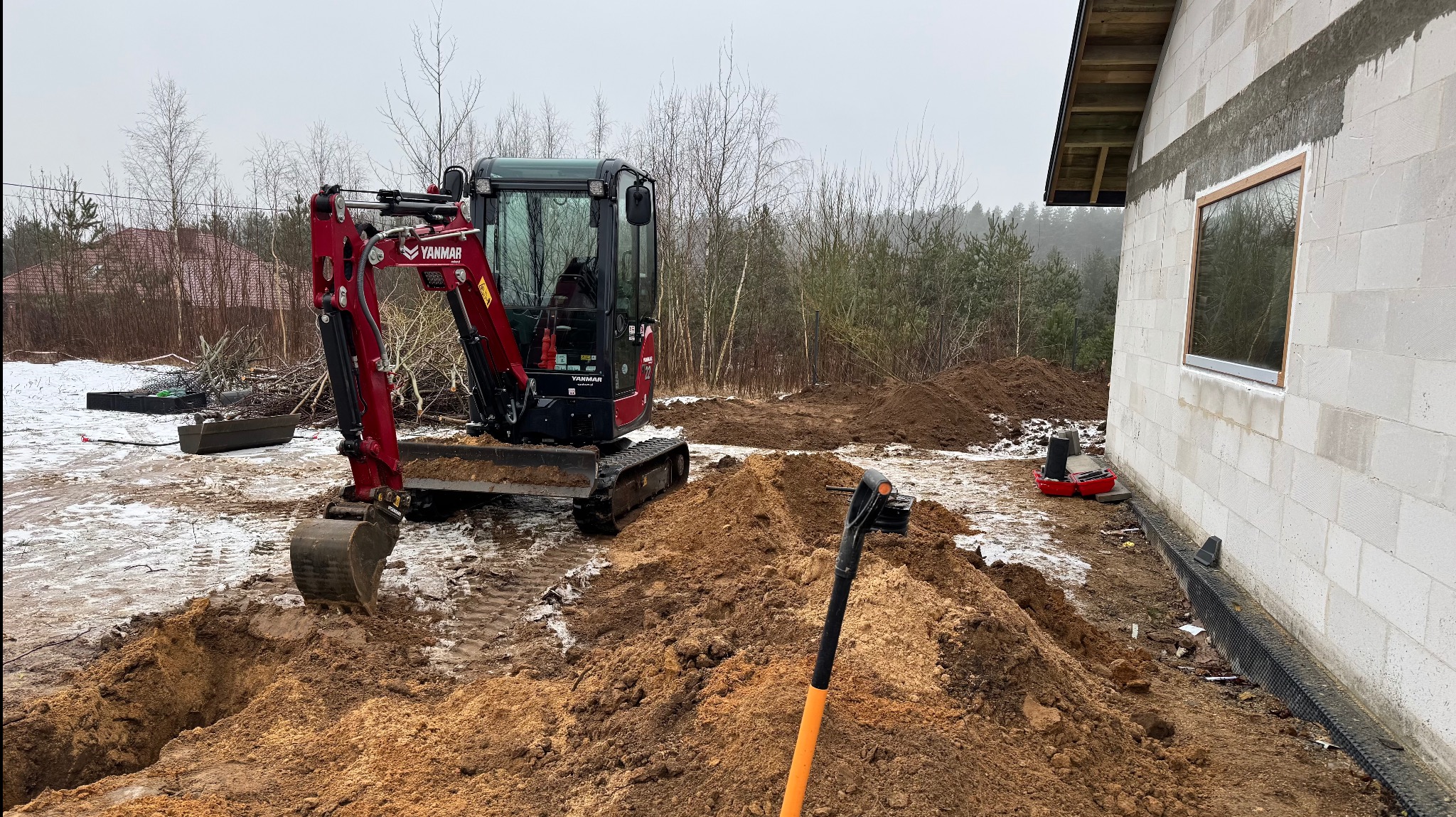 Czerwona minikoparka Yanmar na placu budowy, obok fundamentów domu z bloczków, z widoczną izolacją przeciwwilgociową i resztkami śniegu na ziemi, z łopatą wbita w pryzmę ziemi na pierwszym planie.